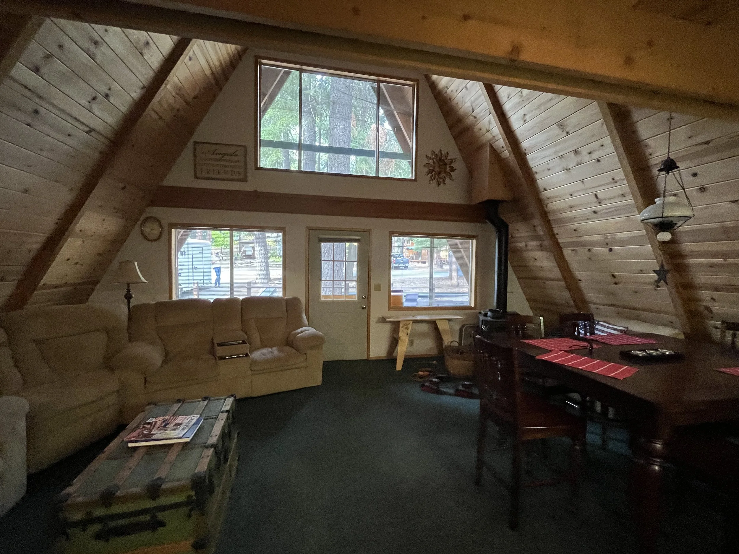 The interior of a cozy cabin living room with wooden walls and ceiling, large windows showing trees outside, a beige sofa, a table with magazines, and a dining table with red placemats.