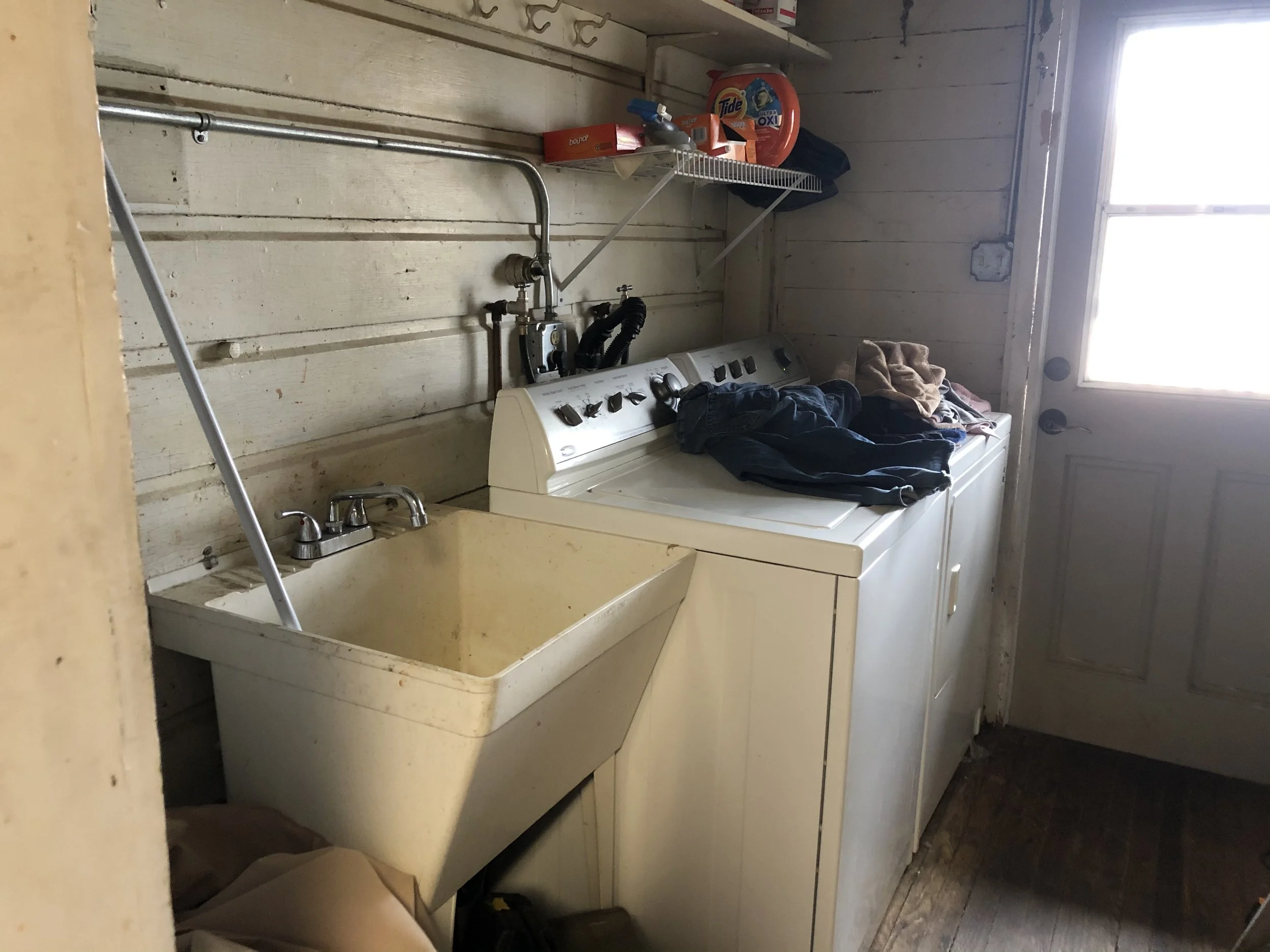 A laundry room with a white washing machine and yellow stained utility sink, above which is a wire shelf holding detergent bottles, with some laundry items on top and a pile of clothes on the washing machine.