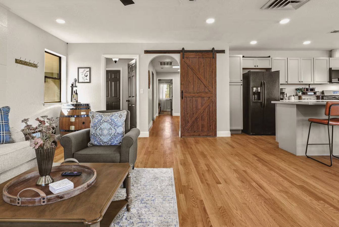 Living room and kitchen area with hardwood floors, wicker couch with plaid pillows, armchair with decorative pillow, coffee table with flowers and remote, wooden sliding barn door, refrigerator, white cabinets, countertop with kitchen appliances and 