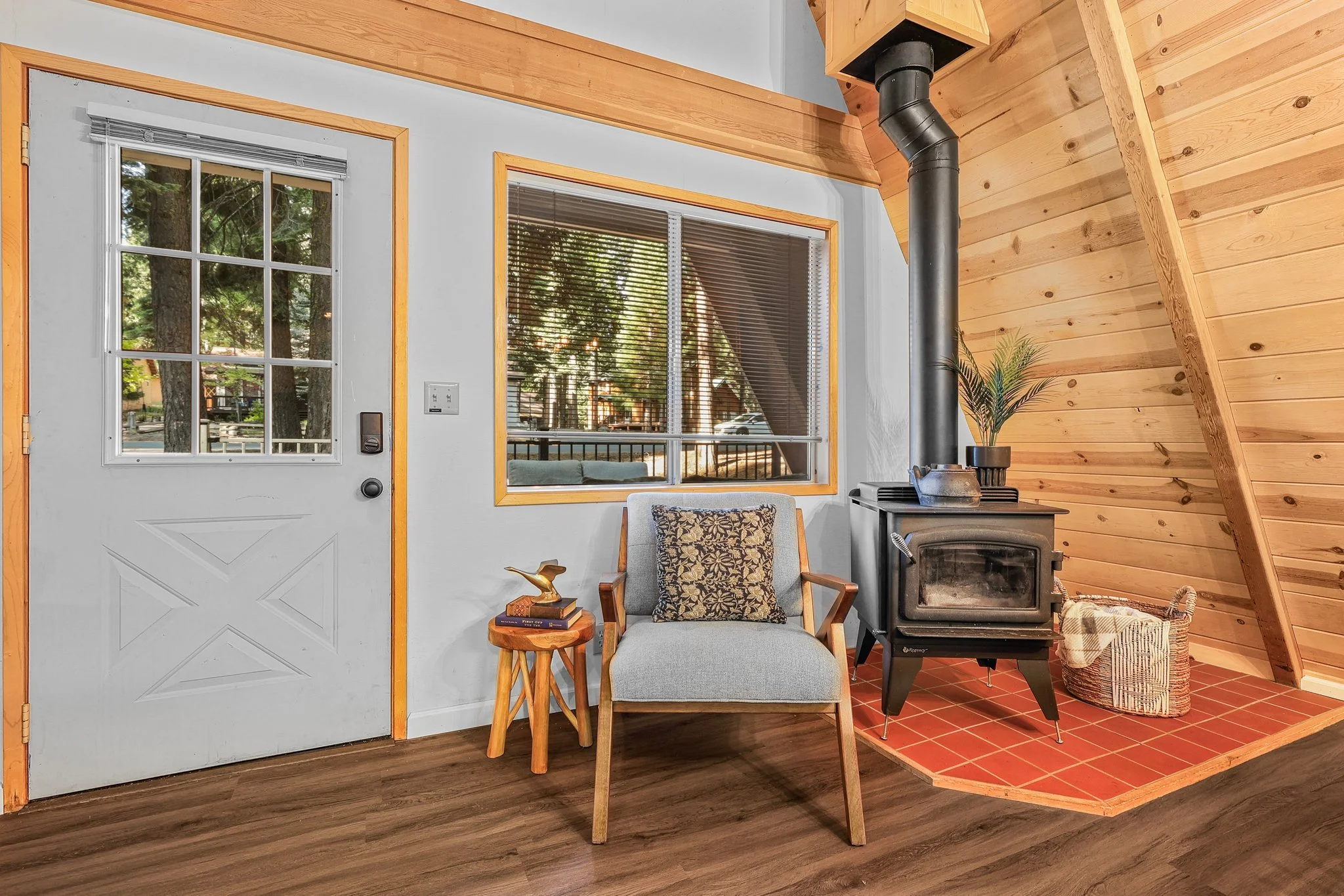 Interior of a cozy room with a wooden and white color scheme. Features include a window with blinds, a door with a window panel, a fireplace with a black pipe, a small wooden side table with books and a decorative bird, a light gray armchair with pat