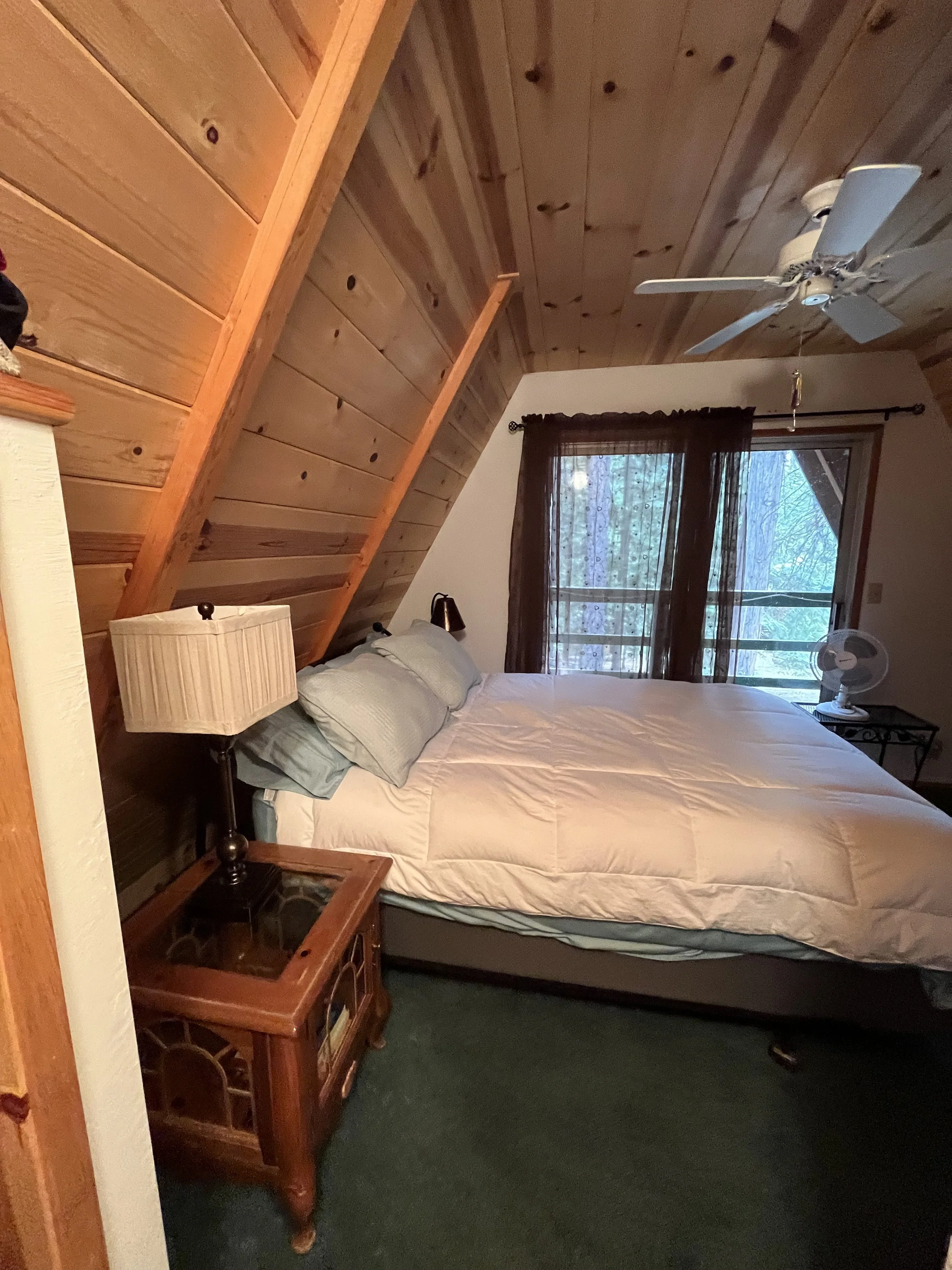 A bedroom with a wooden pitched ceiling, a window with dark curtains, and a bed with light-colored bedding. There are two lamps on bedside tables and a ceiling fan.