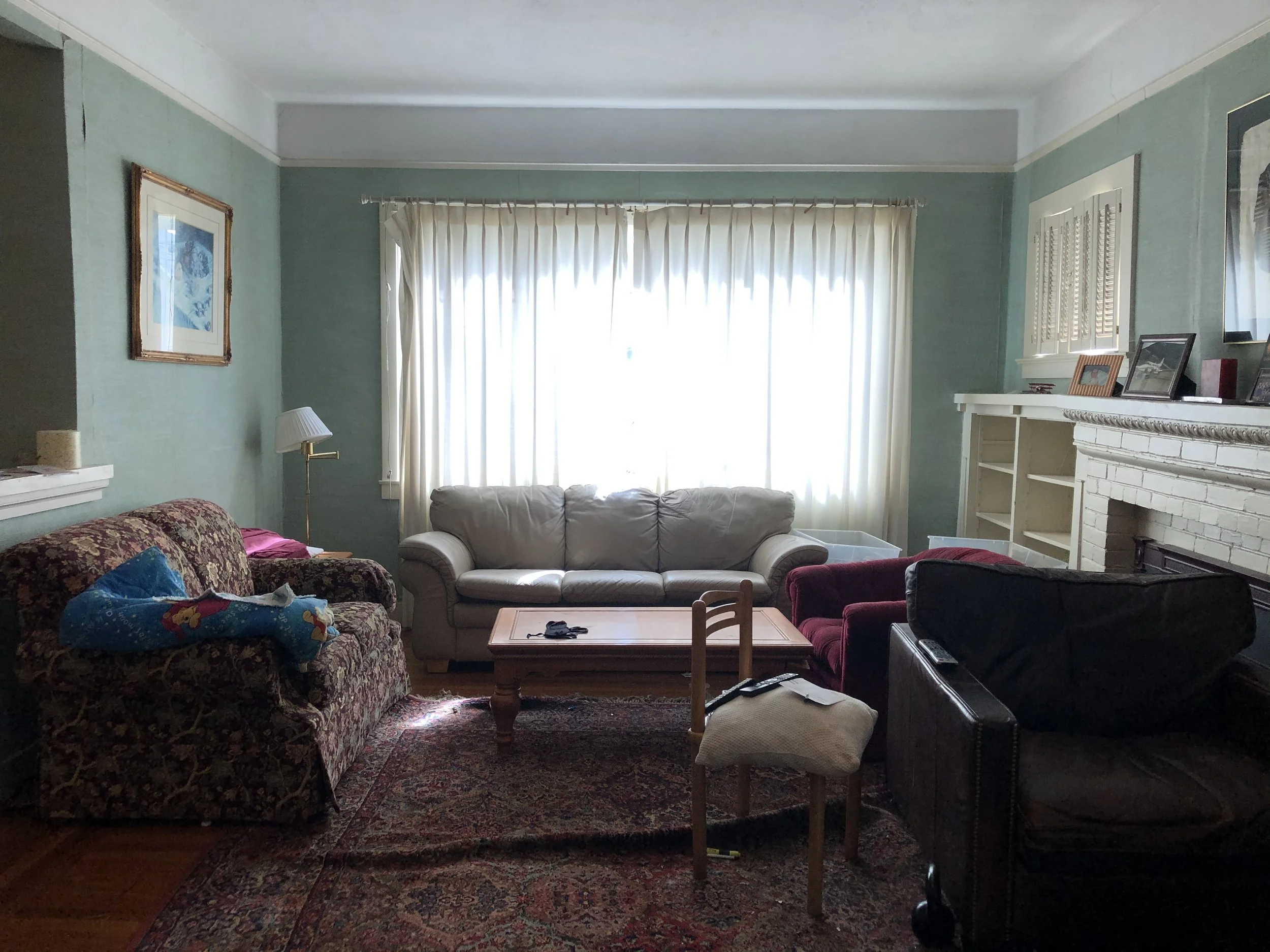 Living room with large window, white curtains, light green walls, and multiple sofas, some with blankets. A small wooden table with objects, a fireplace with picture frames and decorative items, an area rug, and a ceiling with white trim.