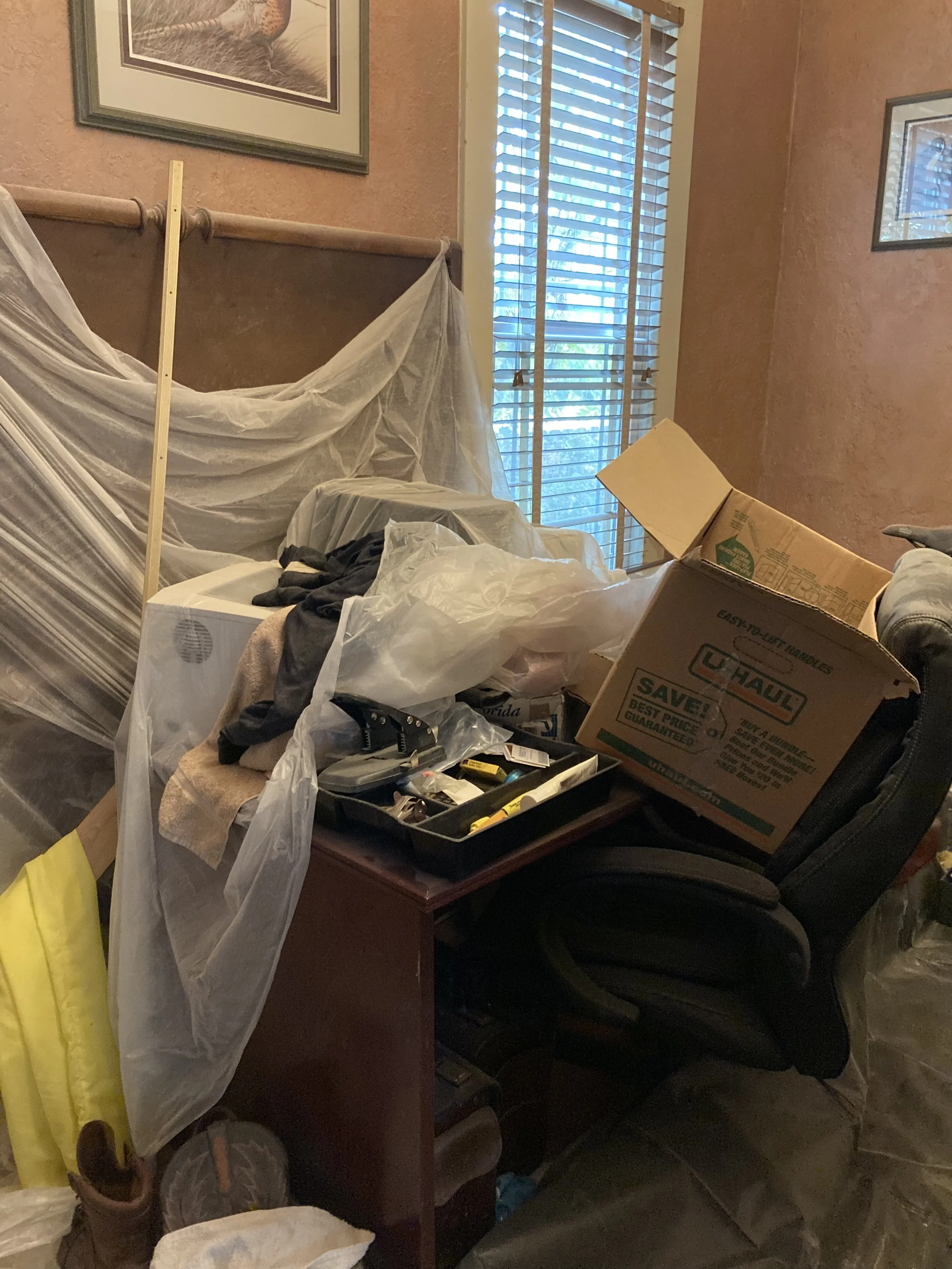 A cluttered room with a bed, a window with blinds, and various boxes and items, including a moving box labeled U-Haul, scattered around.