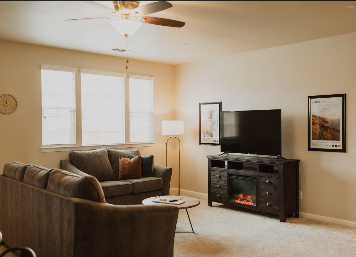 Living room with beige walls, a ceiling fan with lights, a gray sofa with pillows, a round coffee table, a floor lamp, a television on a dark wooden stand, framed pictures on the wall, and a window with blinds.