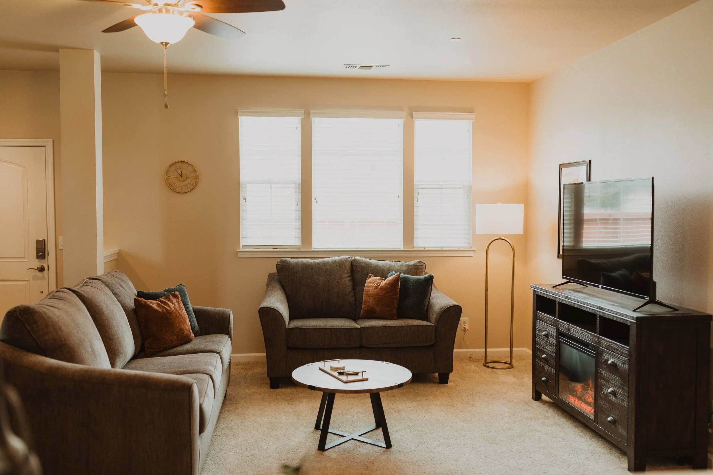 Living room with beige walls, three large windows with white blinds, a ceiling fan, a clock, a gray couch with throw pillows, a side table with a lamp, a round coffee table, and a TV on a black stand with an electric fireplace.