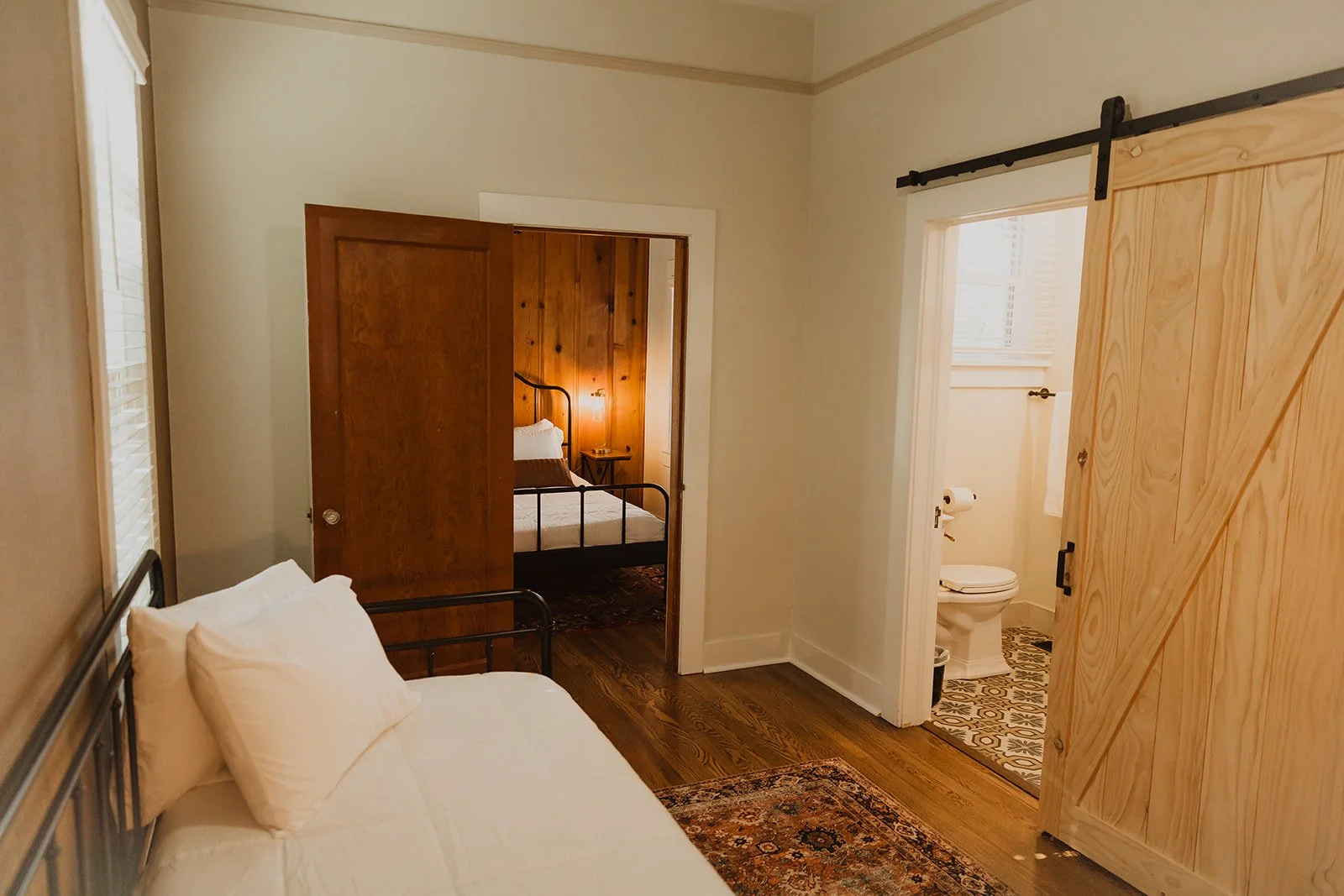 A bedroom with a bed featuring white linens and pillows, a wooden wall with a bed, a sliding barn door, and a bathroom visible through an open door showing a toilet and patterned tile floor.