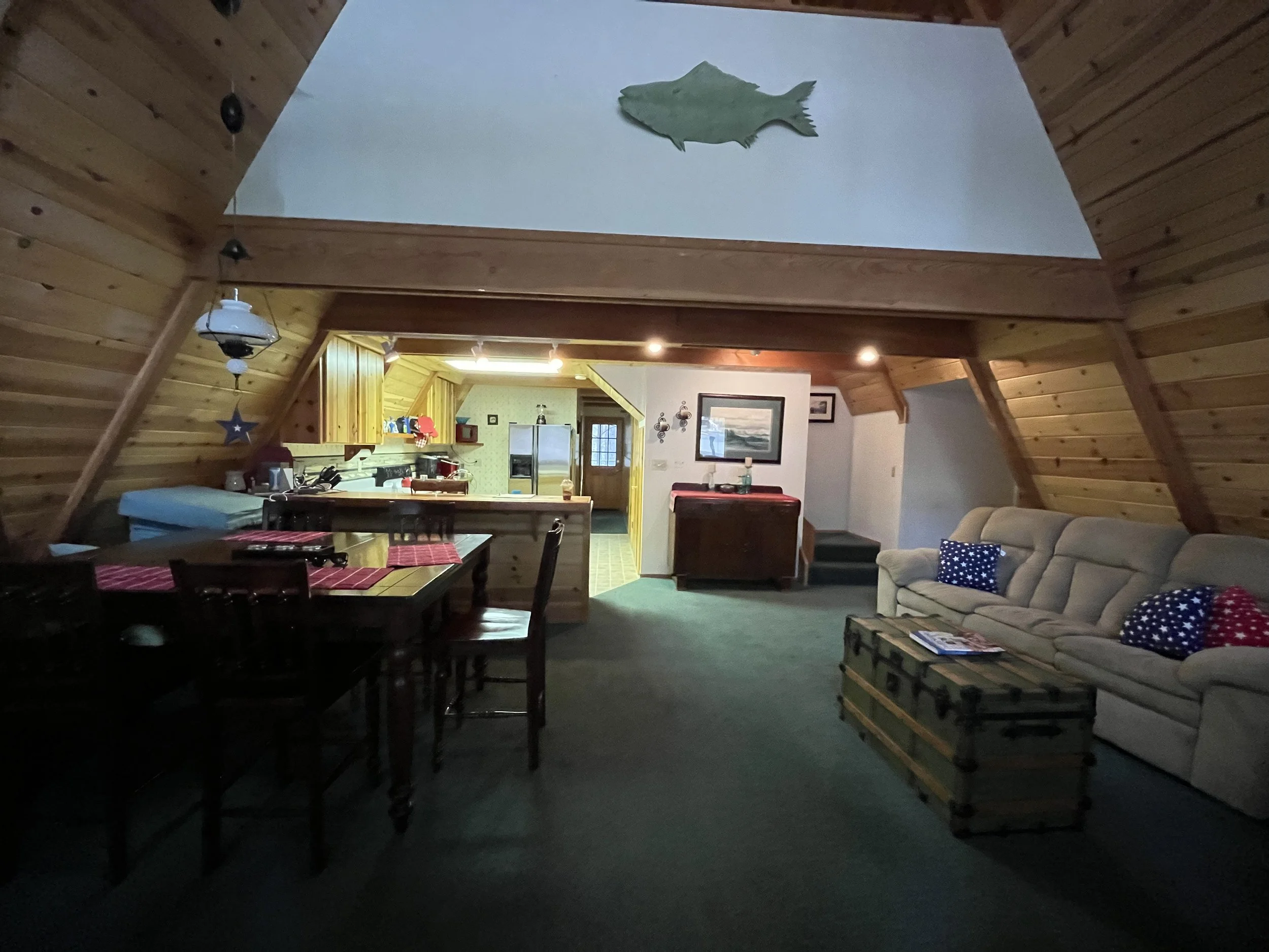 Interior view of a living room and kitchen in a wooden house with sloped ceiling. The living room has a beige sofa with star-patterned pillows, a wooden coffee table with magazines, and a dark green carpet. The kitchen has light wood cabinets with re