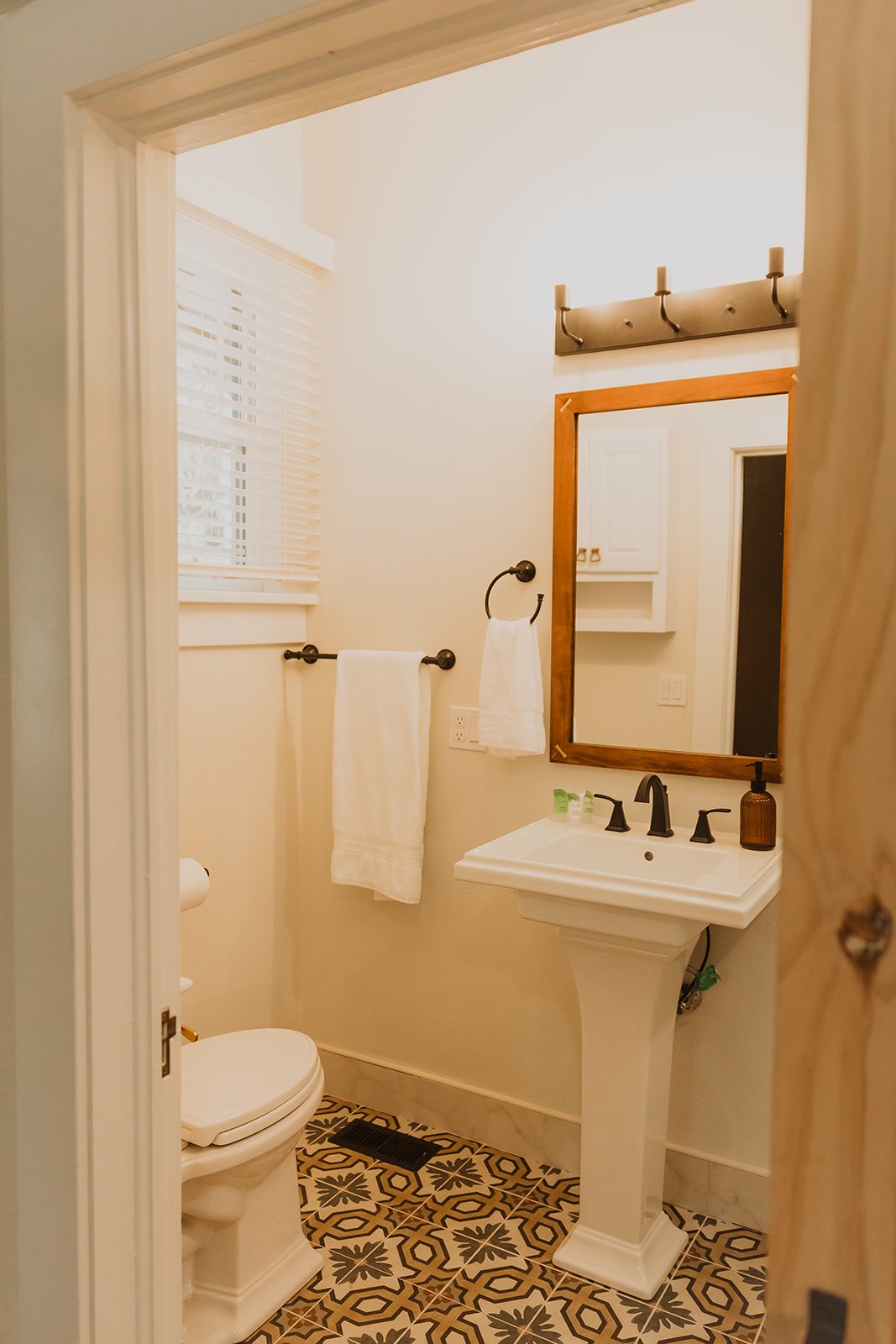 Small bathroom with white walls, featuring a white pedestal sink with black fixtures, a large mirror with a wooden frame, a black towel ring holding a white towel, and a black metal hook strip with four hooks. There is a window with blinds, a white t