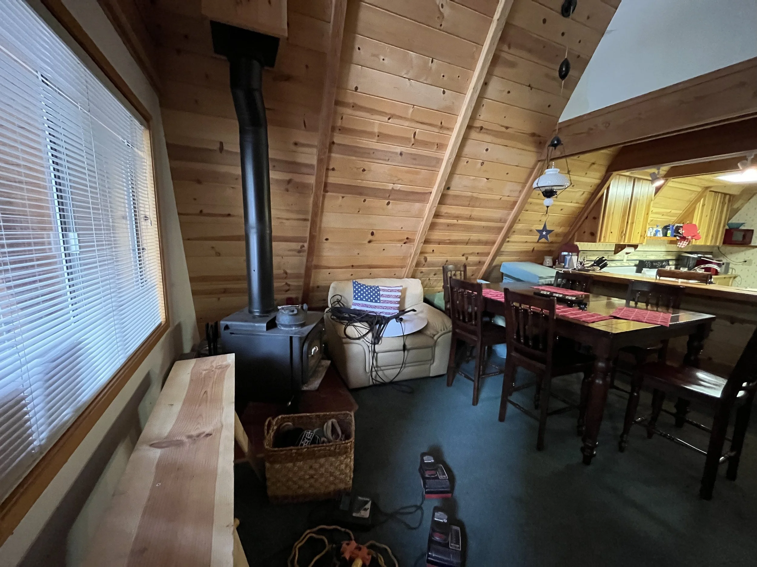The image shows the interior of a room with wood-paneled vaulted ceiling and a large window with blinds. There is a wood-burning stove pipe visible in the middle, and part of a dining area with a table and chairs with red checkered placemats. The roo