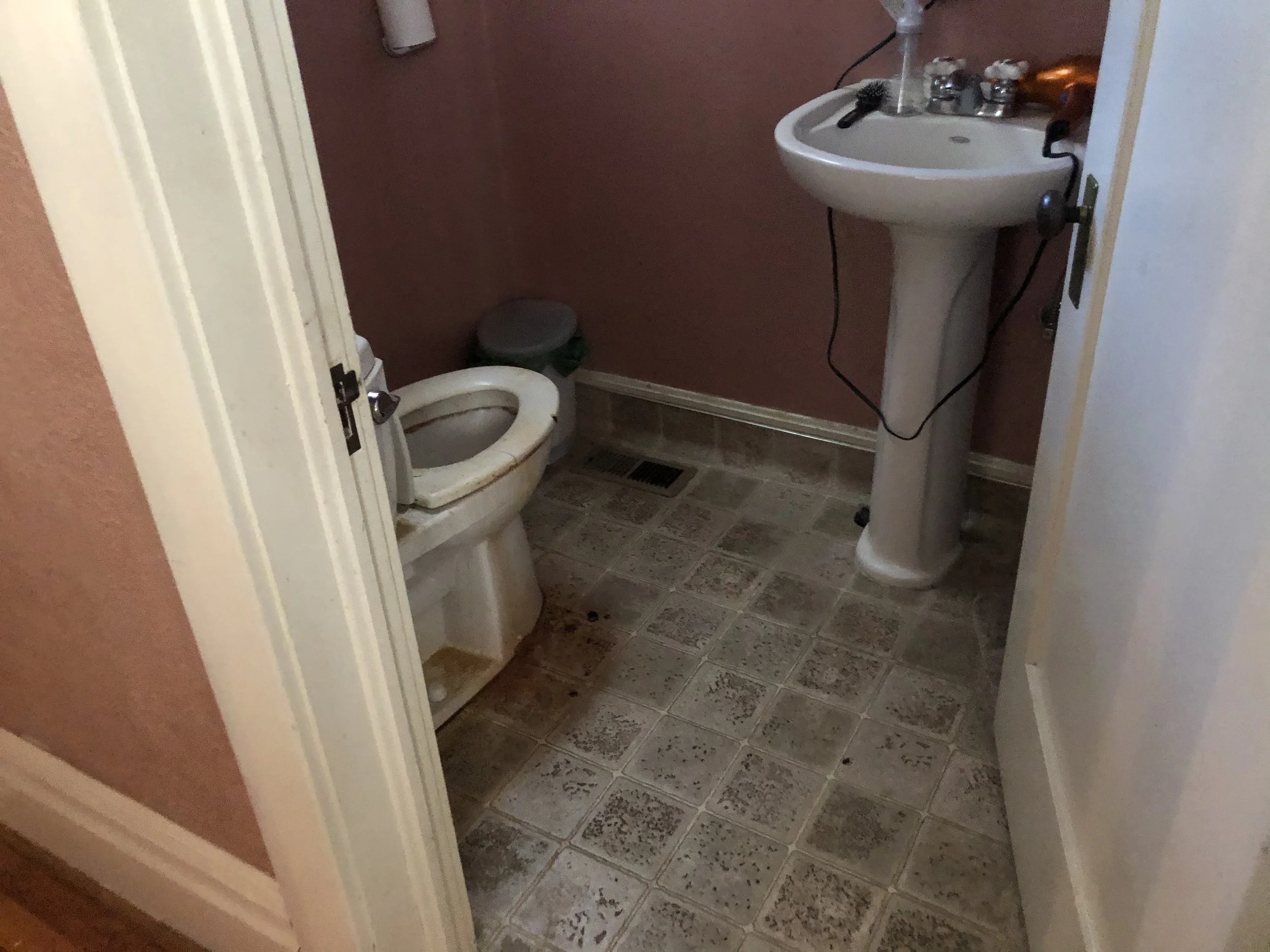 A small bathroom with a white toilet, a white pedestal sink, and brown painted walls. Various toiletries are on the sink, and there is a black hairbrush next to them. The floor has patterned tiles, and there is a vent below the toilet.