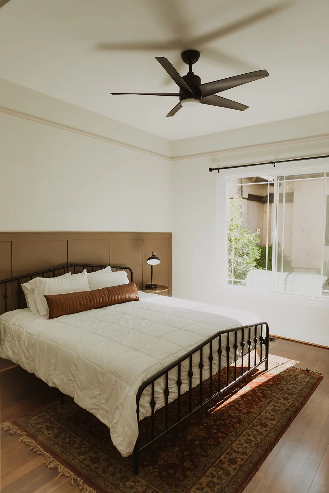A bedroom with a black metal bed frame, white bedding, a brown bolster pillow, a small black nightstand with a lamp, a ceiling fan, a large window with white curtains, and a patterned rug on wooden floors.