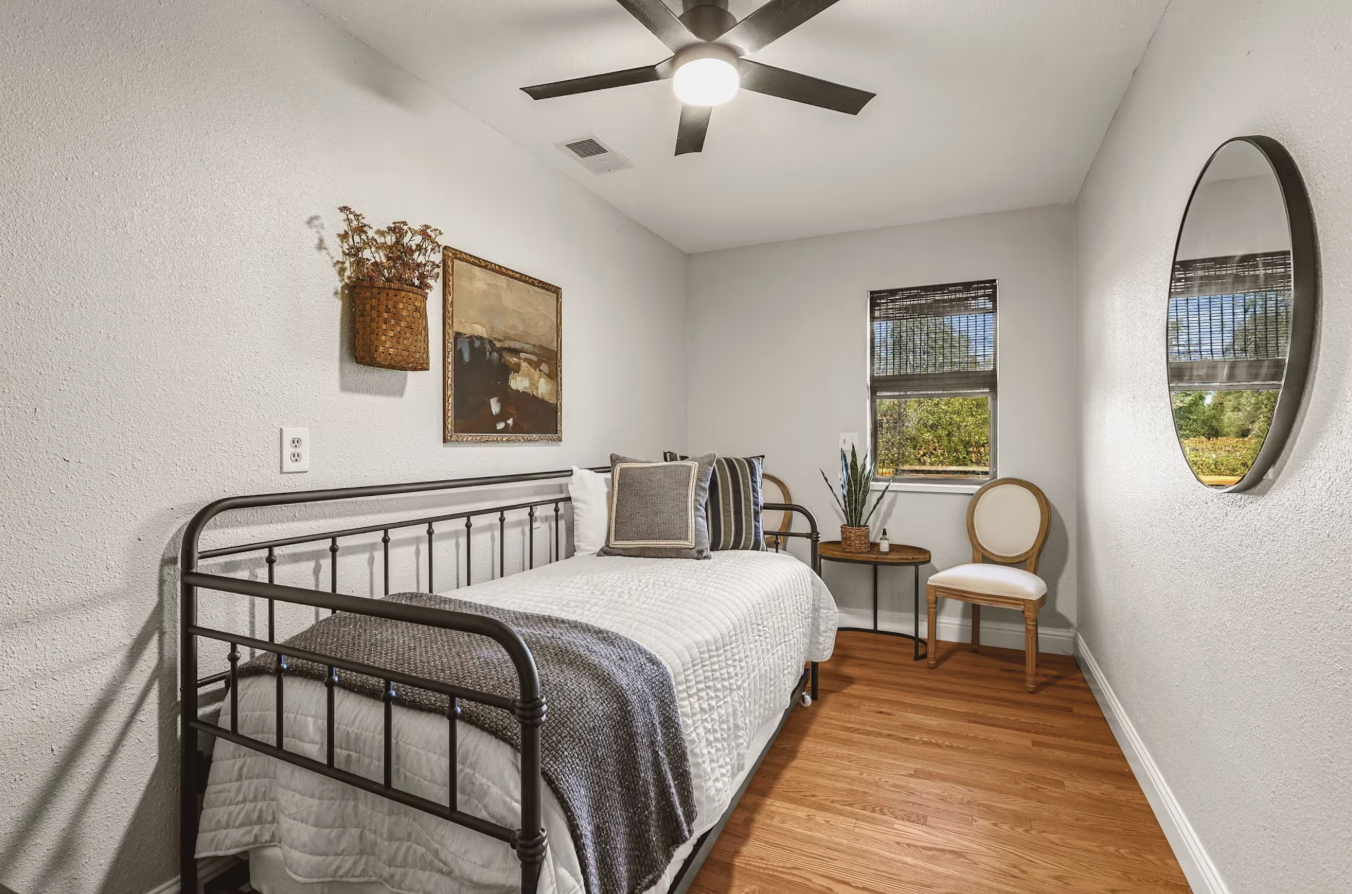 A cozy bedroom with a metal bed frame, white bedding, and decorative pillows. A small wall-mounted basket with dried flowers, a framed artwork, a wooden chair, and a window with blinds are visible, along with a ceiling fan and hardwood floors.