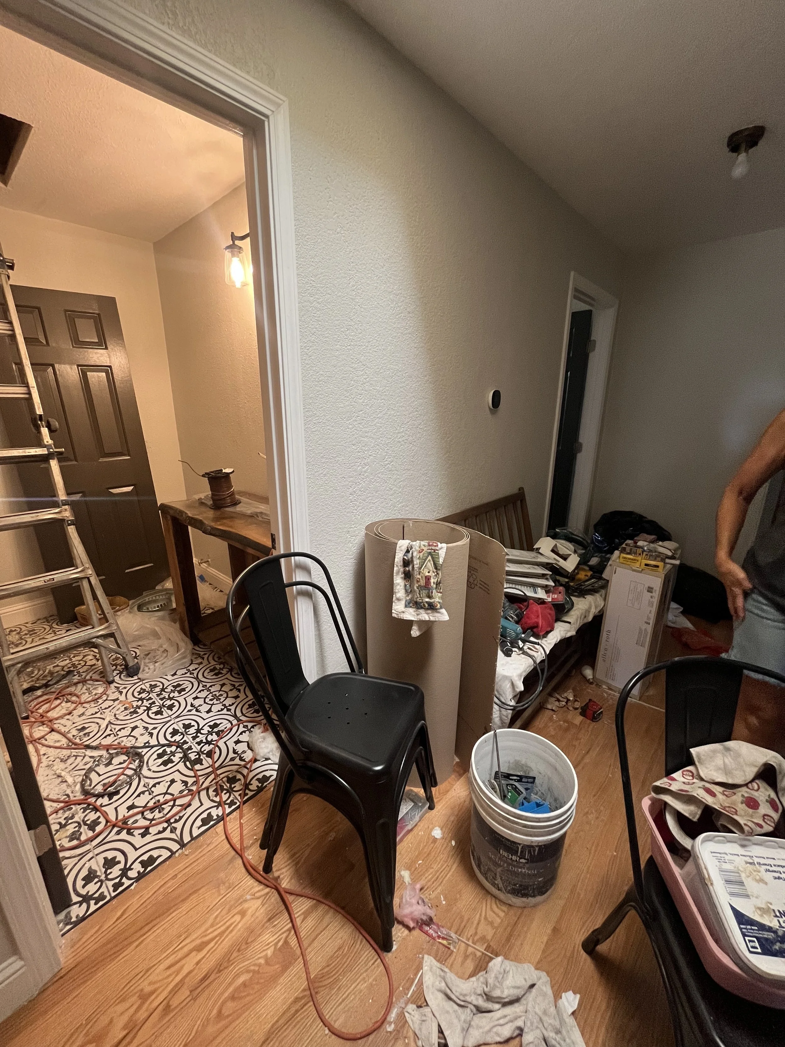 Partially cleared cluttered room with chairs, a bucket, tools, and construction materials, with a doorway leading to a small area with black door and patterned tile floor.
