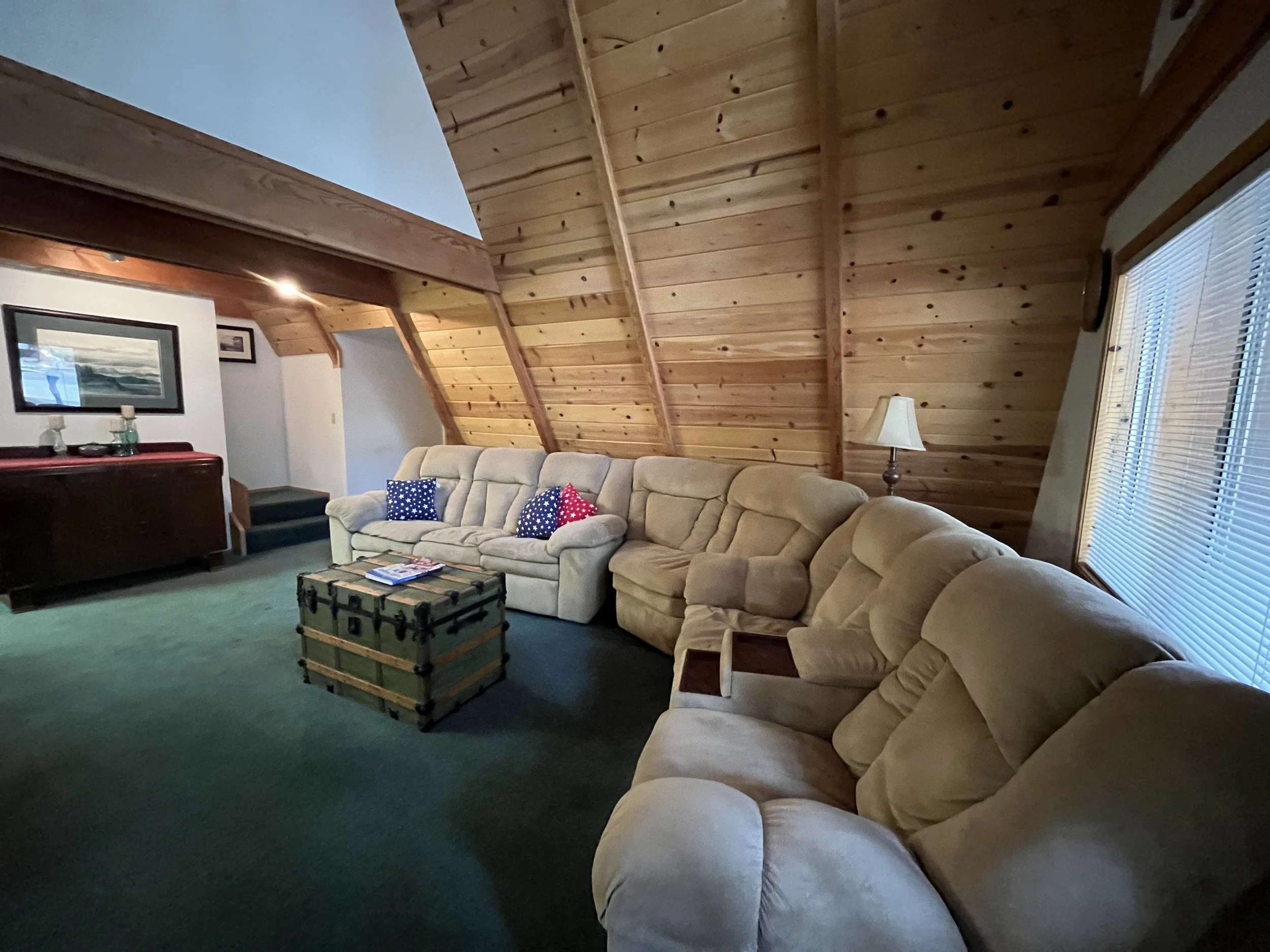 Living room with a large beige sectional sofa, pillows with patriotic colors, wooden vaulted ceiling, window with blinds, side lamp, and framed art on the wall.