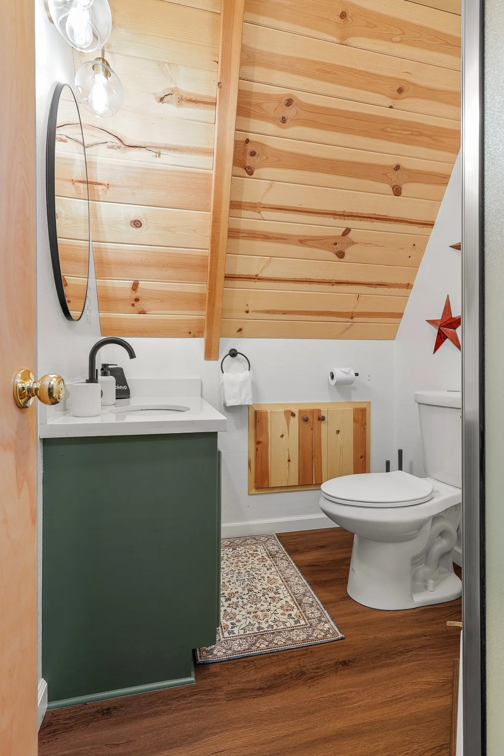 Small bathroom with a sloped wooden ceiling, white walls, and wood accents. Contains a white toilet, a green vanity with a white countertop and black faucet, an oval mirror, decorative glass light fixtures, a wall-mounted wooden cabinet, a patterned 