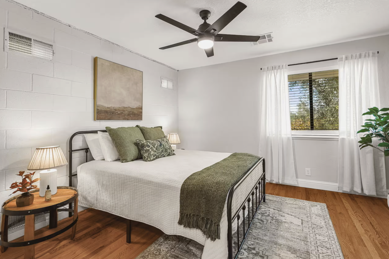 A bedroom with a white wall, a metal bed frame, white bedding, green pillows, a bed runner, and a painting above the bed. There are two lamps on a round wooden side table, a window with white curtains, a plant in the corner, and a ceiling fan.