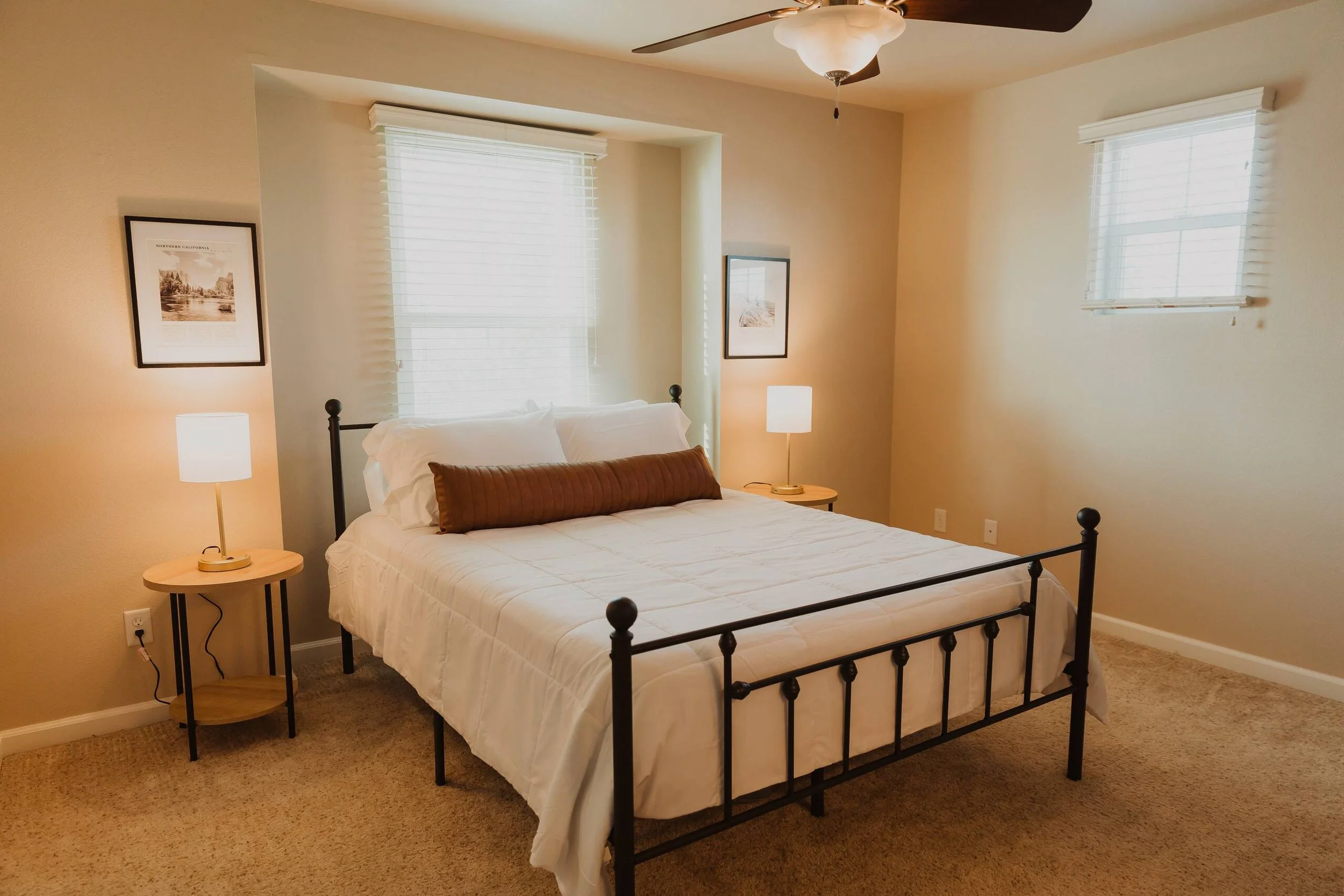 A bedroom with beige walls, a black metal bed frame with white bedding and a brown pillow. There are two windows with white blinds, two framed pictures on the walls, and two small side tables each with a white lamp.
