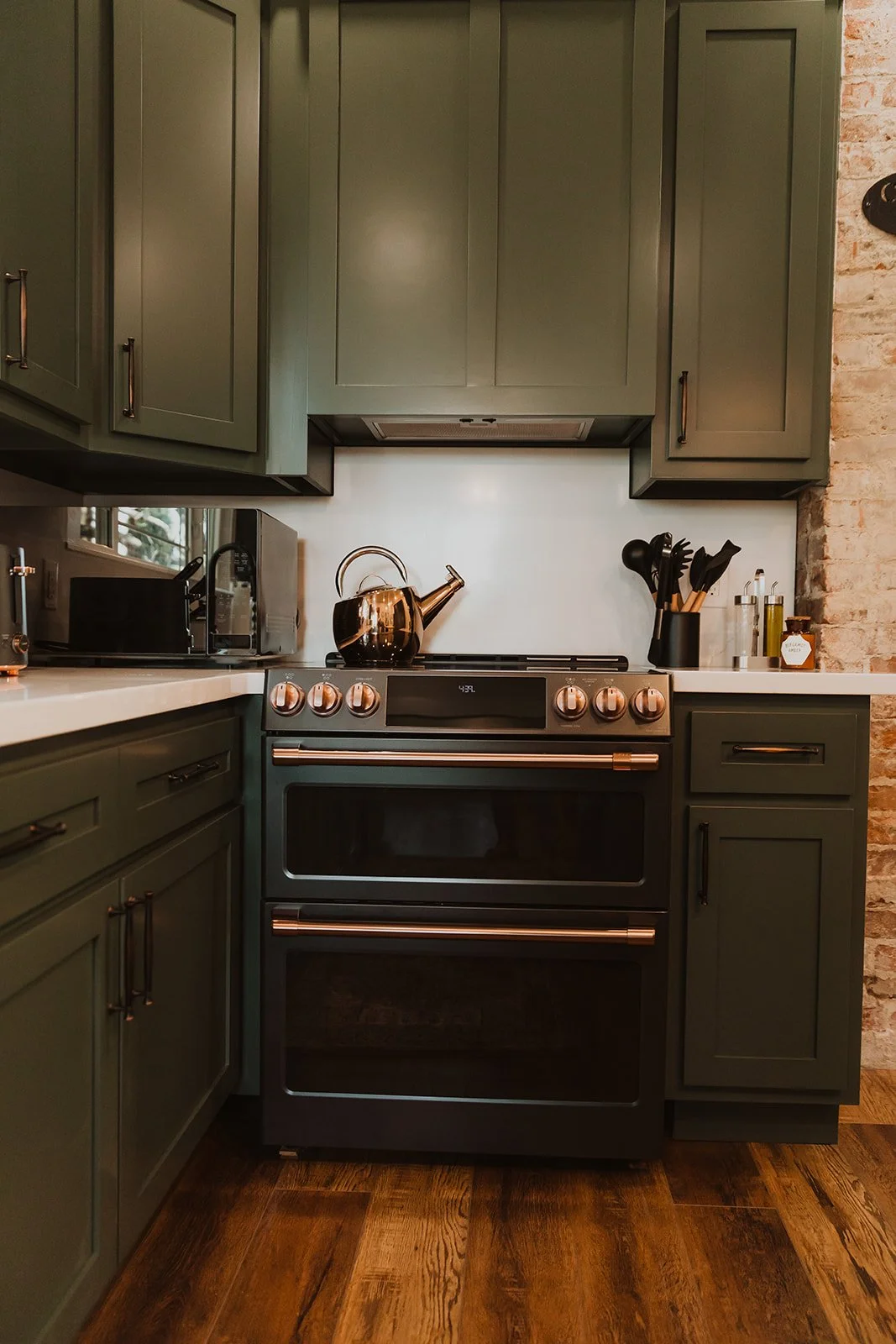 Kitchen with dark green cabinets, a black stove with copper accents, a kettle on the stove, and various kitchen tools and containers on the countertops.