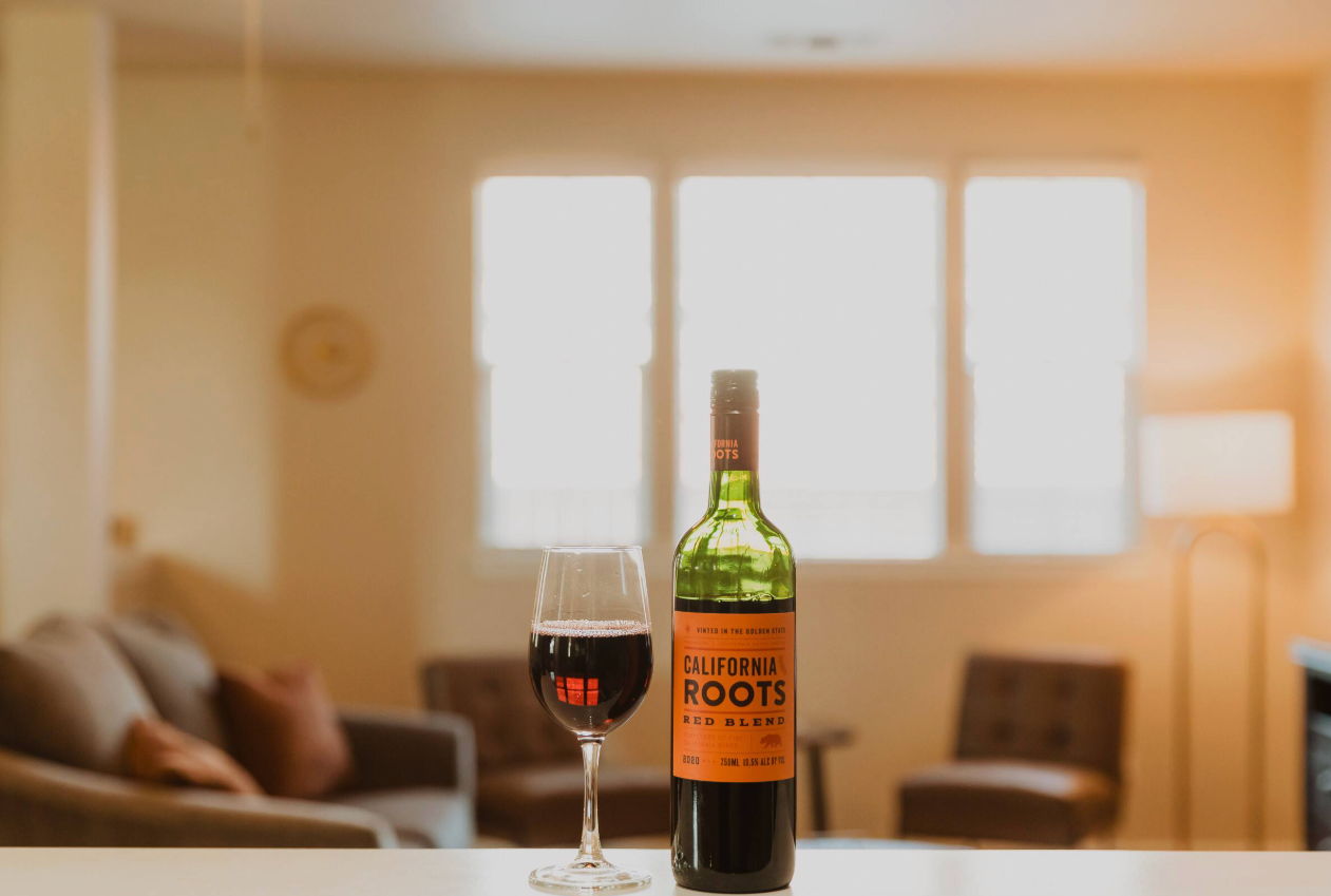 A bottle of California Roots red blend wine and a glass of red wine on a white countertop in a living room with beige walls, brown sofas, and large windows letting in sunlight.