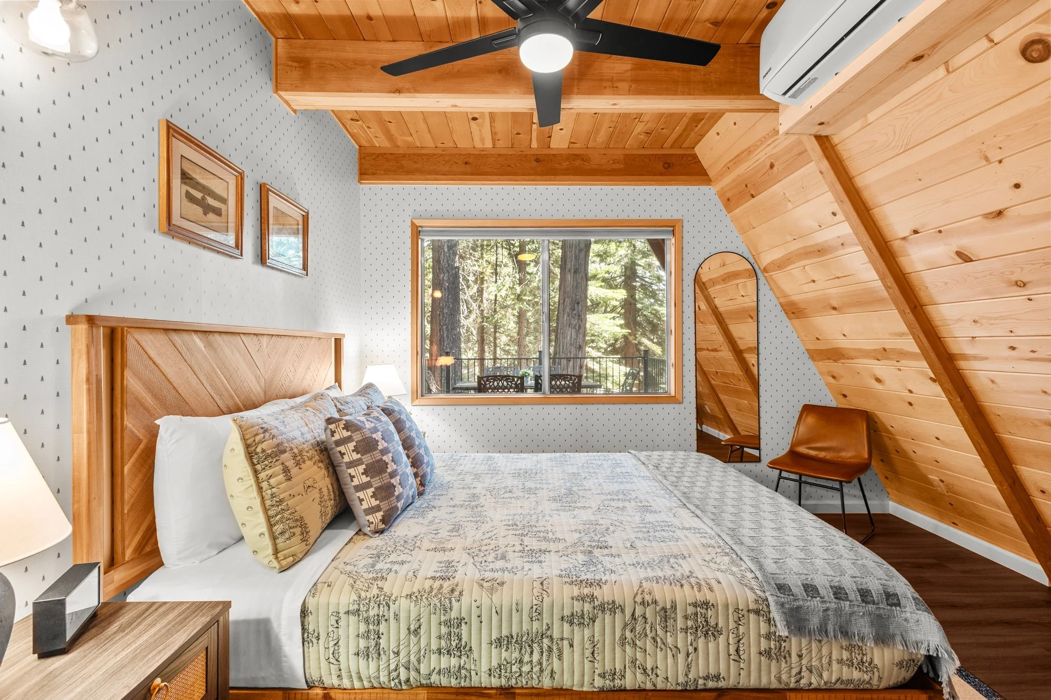 A cozy bedroom with a large bed with patterned bedding and pillows, wooden headboard, and bedside table with a lamp. The room has a window showing a forest view, wood-paneled ceiling with a ceiling fan, and framed artwork on the wall. There is also a