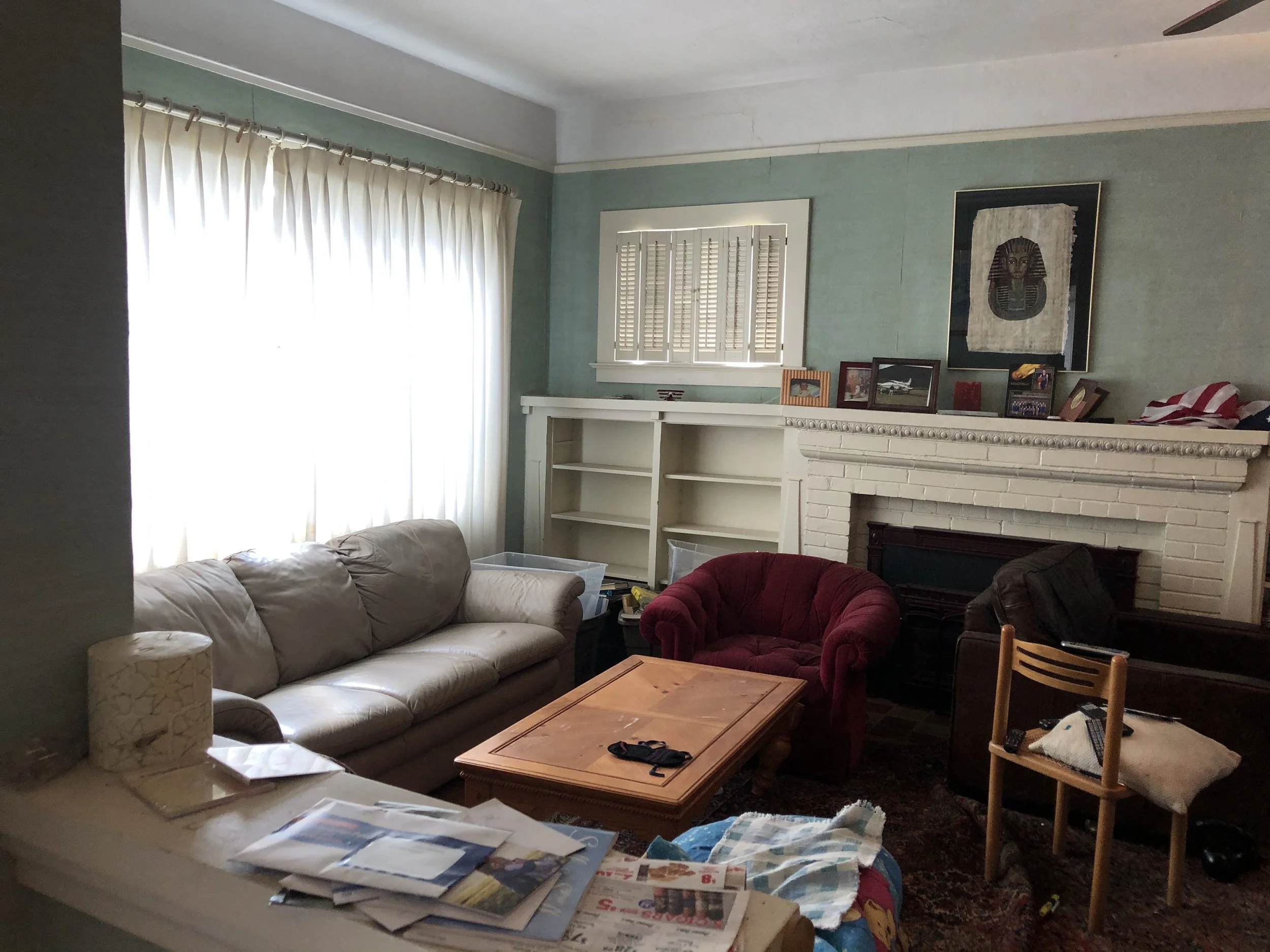 A cluttered living room with a beige couch, red armchair, wooden coffee table, and scattered magazines and papers. The room has a large window with white curtains, a white fireplace mantel with various framed photos, and a piece of wall art featuring