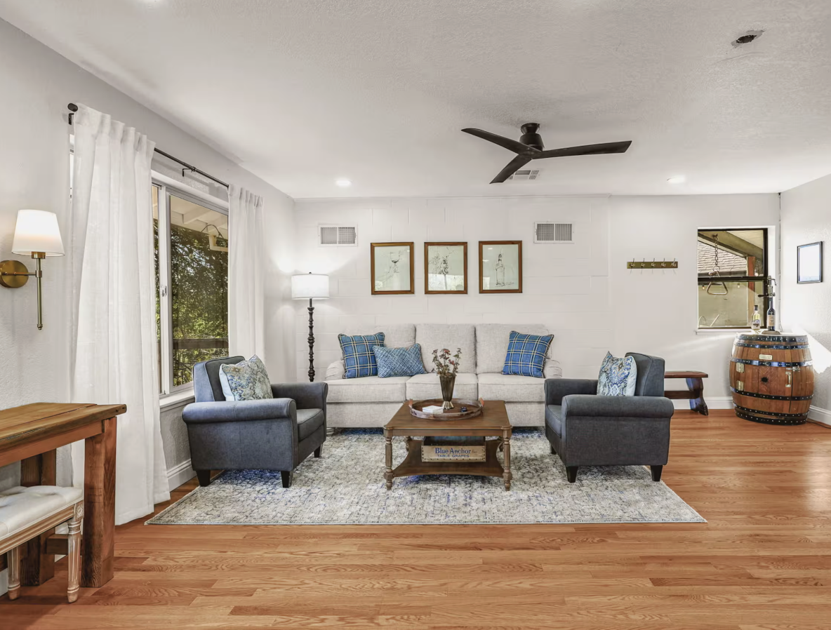 Living room with white walls, hardwood floors, a white sofa with blue plaid pillows, two gray armchairs with patterned pillows, a wooden coffee table, a patterned area rug, and a barrel-shaped wine barrel in the corner. There are wall-mounted and flo