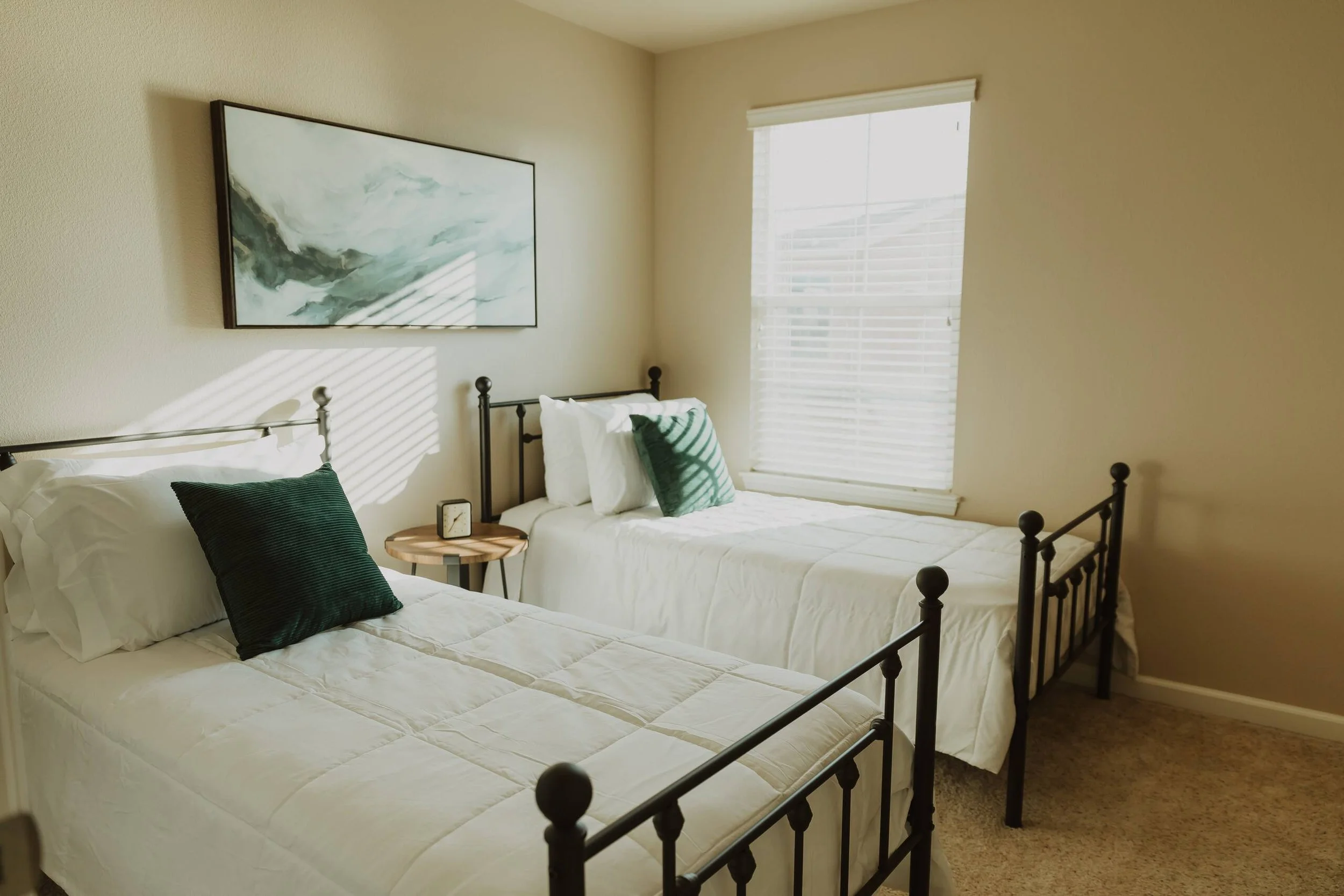 Bedroom with two beds, black metal bed frames, white bedding, green pillows, a small round wooden nightstand, a small clock, and a large window with white blinds. Art painting hanging on the wall.