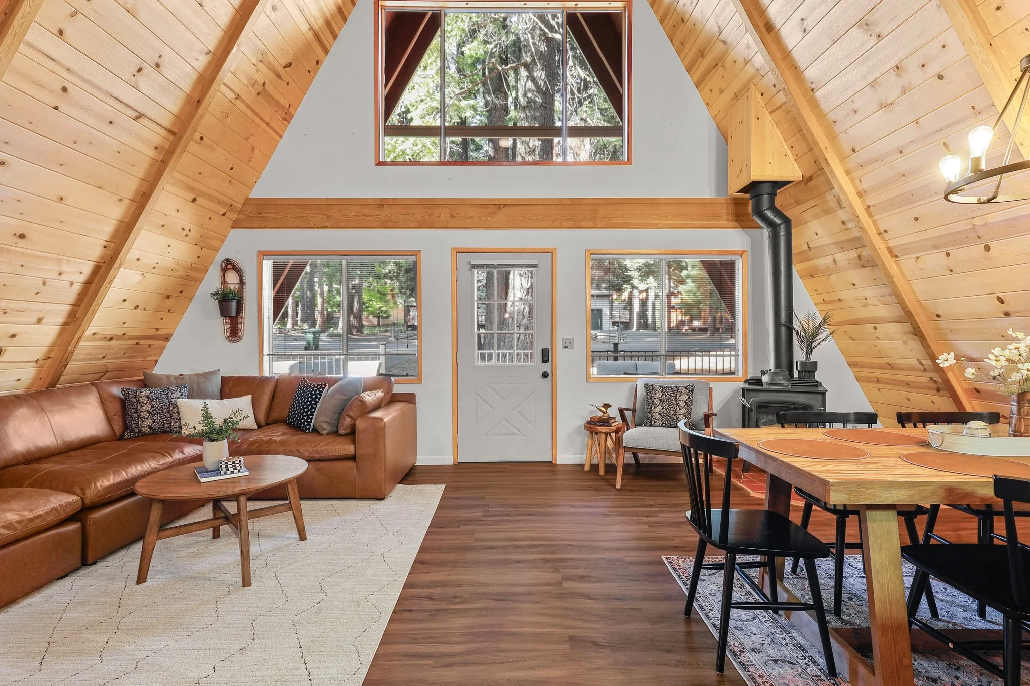 Living room with wooden vaulted ceiling, large windows, leather sofa, fireplace, dining table, and wood flooring.