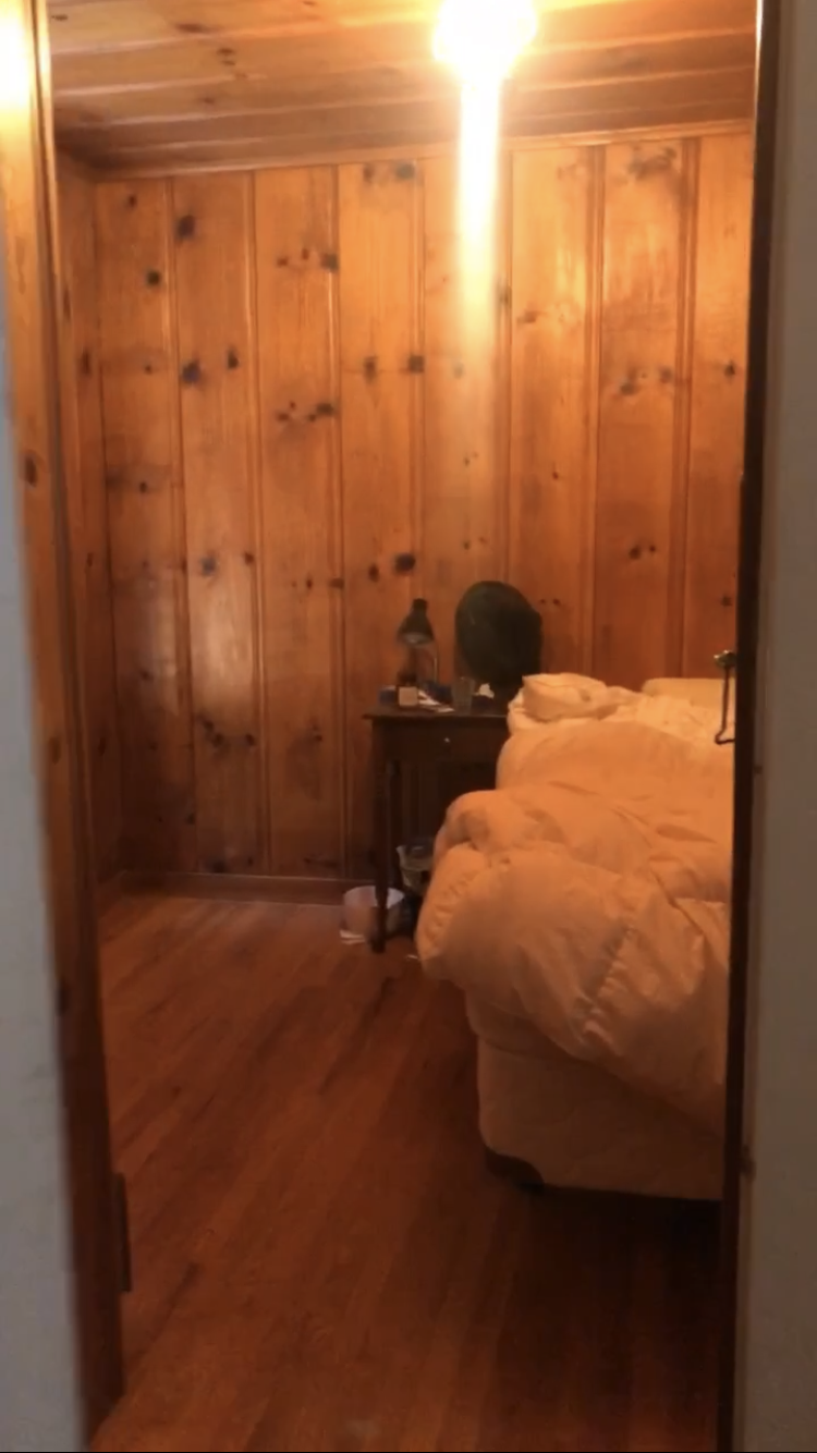 A cozy room with wood-paneled walls, a bed with an orange or beige comforter, a wooden nightstand with a lamp, and a warm light overhead.