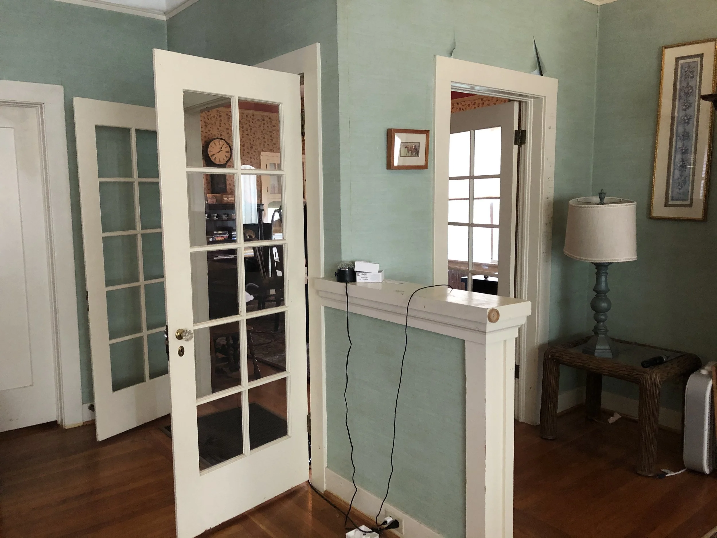 Part of an interior room with green painted walls, a glass-paneled door open to another room with floral wallpaper, a framed picture of horses on the wall, a tall table lamp with a decorative base, a wicker chair, and some electrical cords and boxes 