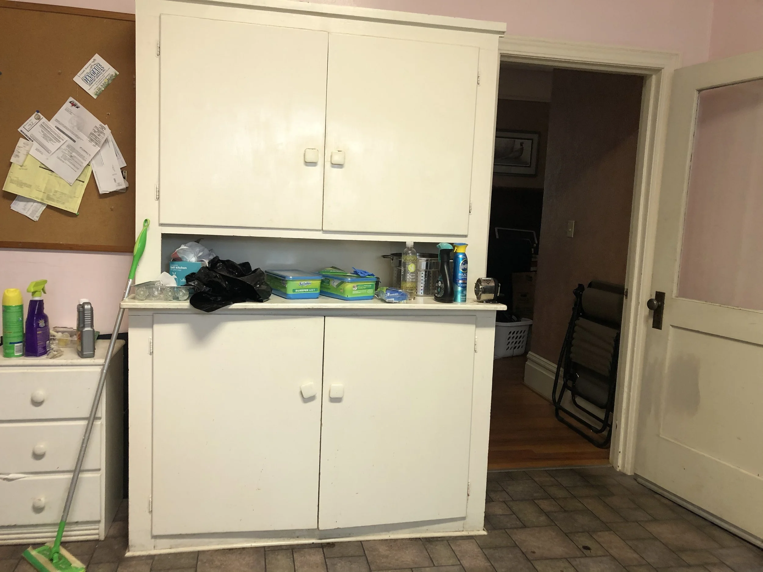 A small kitchen with a white cabinet and a bulletin board with papers on the wall. The countertop has cleaning supplies, a plastic bag, and some green detergent boxes. There are folding chairs stacked in the corner near an open door leading to anothe