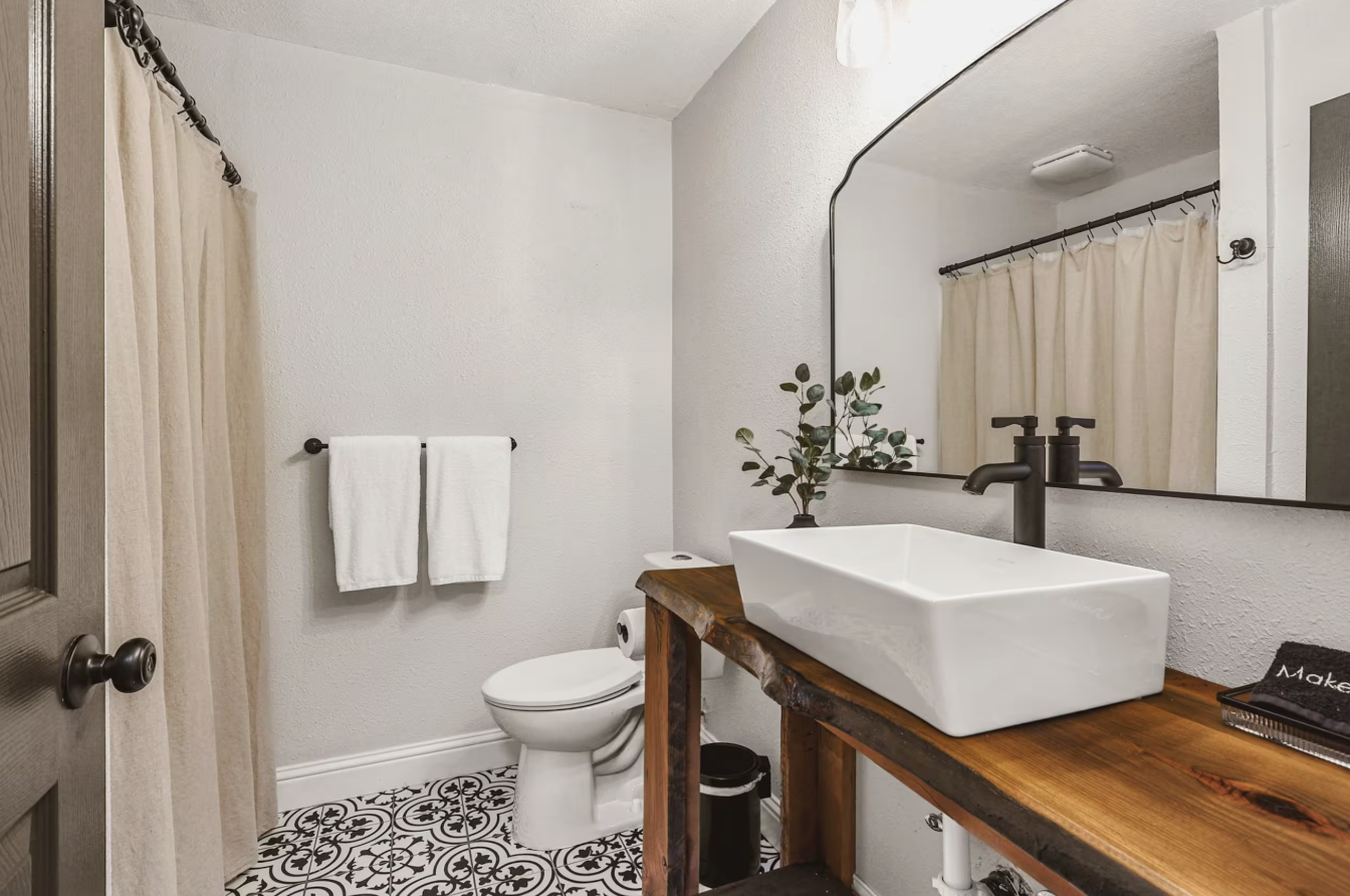 Modern bathroom with white walls, black and white decorative floor tiles, a wooden vanity with a rectangular vessel sink, a black faucet, a large mirror, a potted plant, a toilet, beige curtains on a rod, and white towels on a towel rack.