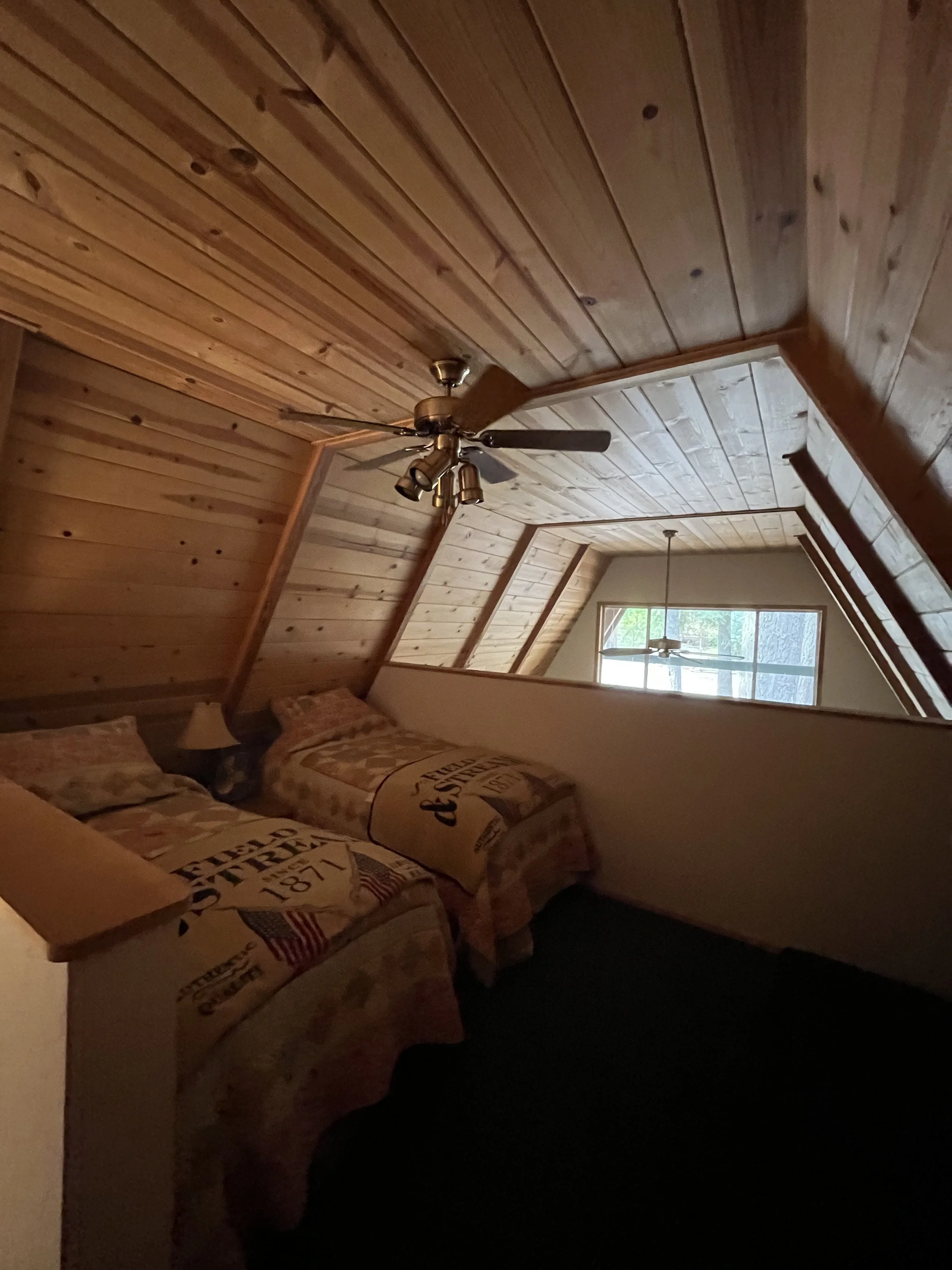 A bedroom with wooden paneled ceiling and walls, a window letting in natural light, and bed with American-themed bedding, including pillows with stars and stripes.