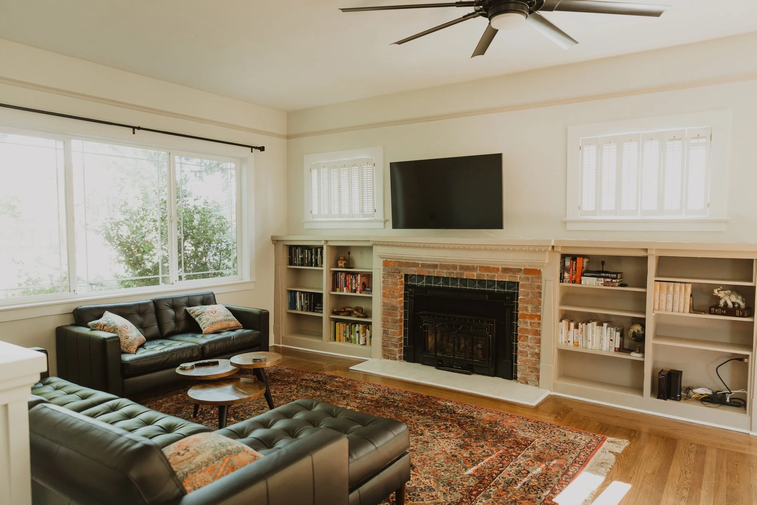 Living room with large windows, black leather sofa, wooden coffee tables, brick fireplace, built-in bookshelves, and wall-mounted TV.