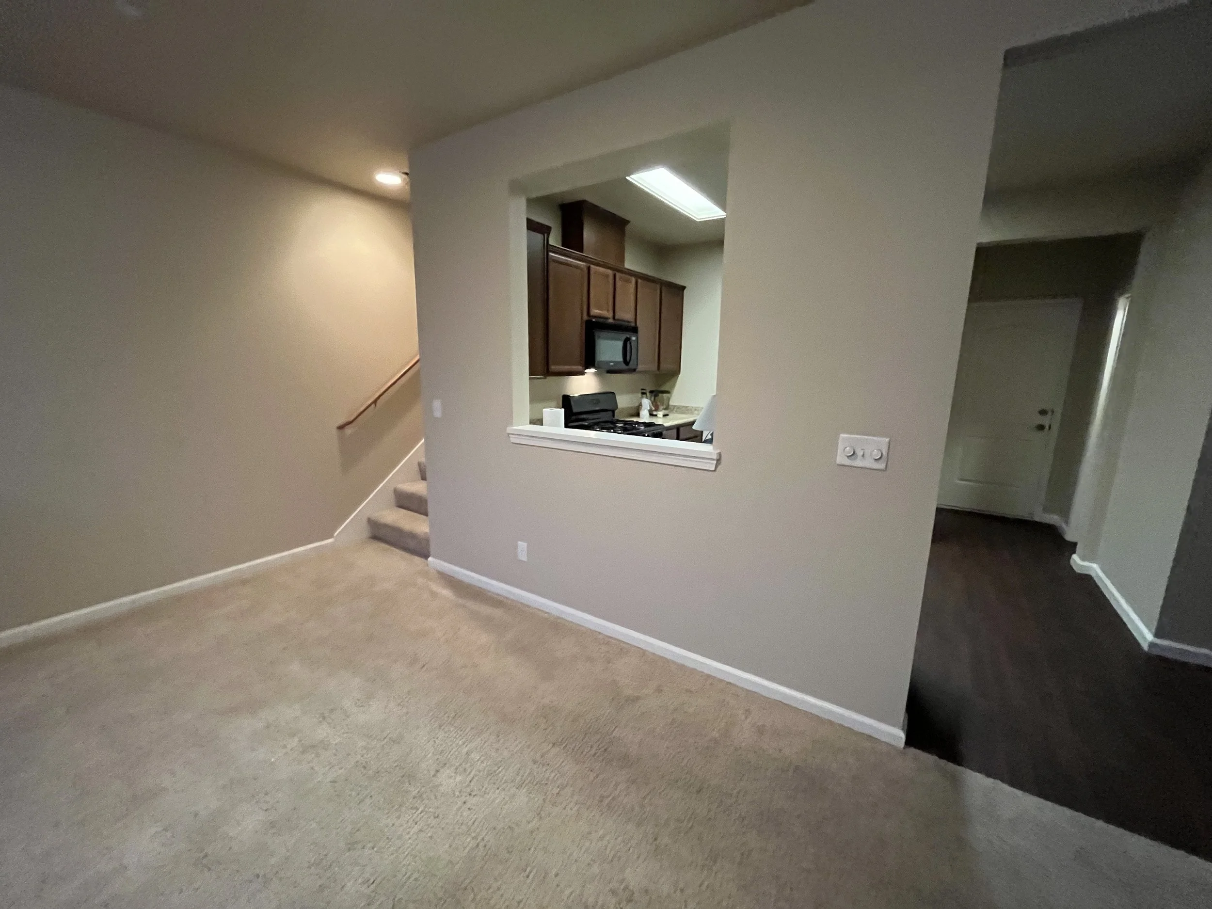 Living room with beige carpet, beige walls, and a staircase. Kitchen area with wooden cabinets and a stove is visible through a partial wall opening.