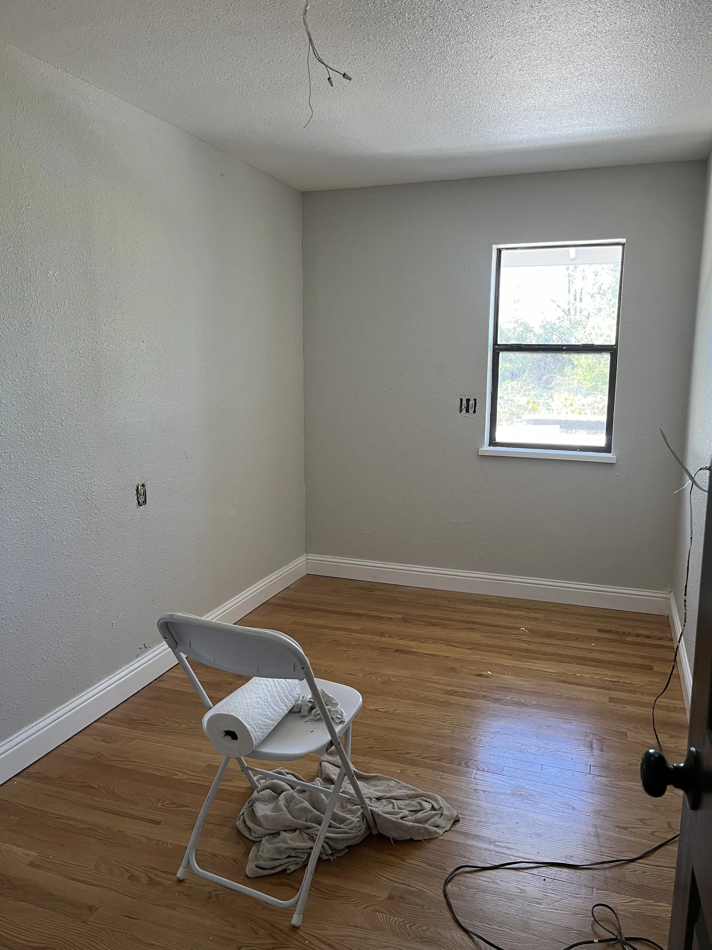 A small empty room with light-colored textured walls, hardwood floors, a window on the right wall, and a foldable chair with a paper towel roll, cloth, and some tools on it, indicating ongoing renovation or painting.