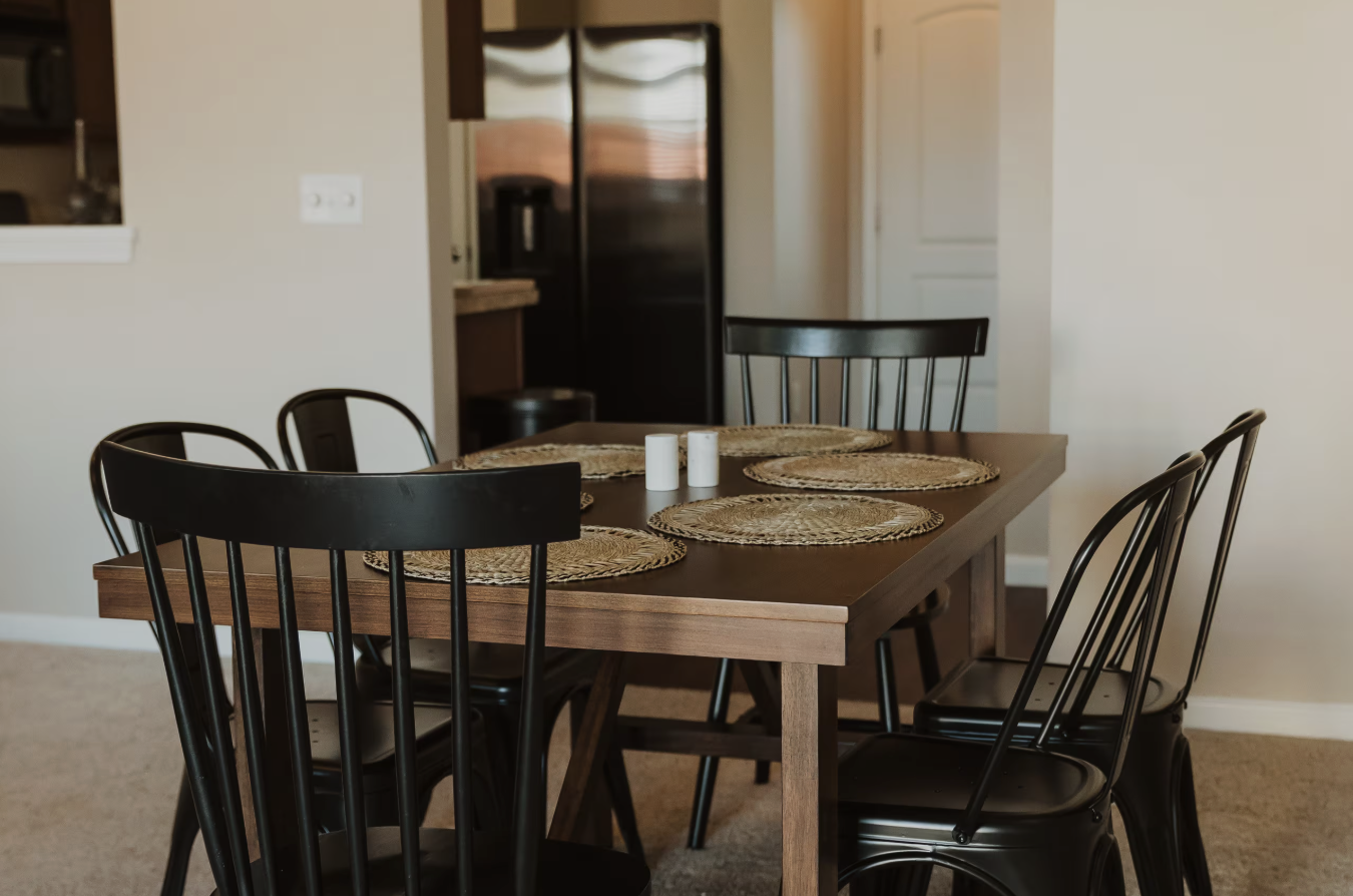 A wooden dining table with five black chairs around it, set with four woven placemats and two salt shakers, in a dining area near a kitchen with a black refrigerator.