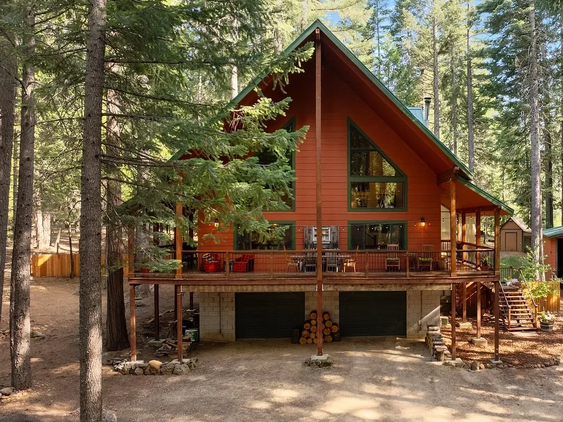 A two-story wooden cabin with a steep green roof, large triangular windows, and a spacious deck in a forested area surrounded by tall trees.