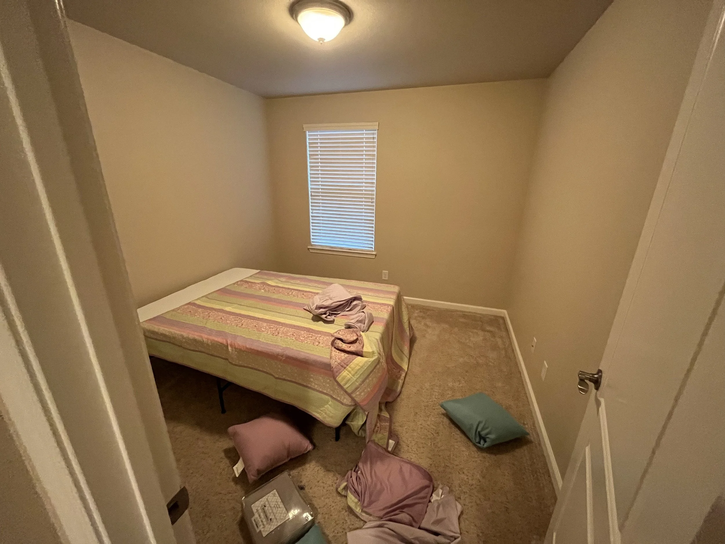 A small, sparsely decorated bedroom with beige walls, a window with blinds, a bed with a colorful quilt, and clothes and items scattered on the floor.