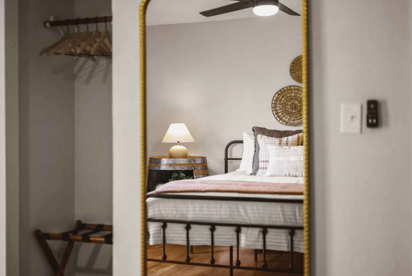 Reflective mirror showing a bedroom with a metal bed frame, a nightstand with a lamp, decorative wall hangings, and an open closet with hanging space and a luggage stand.