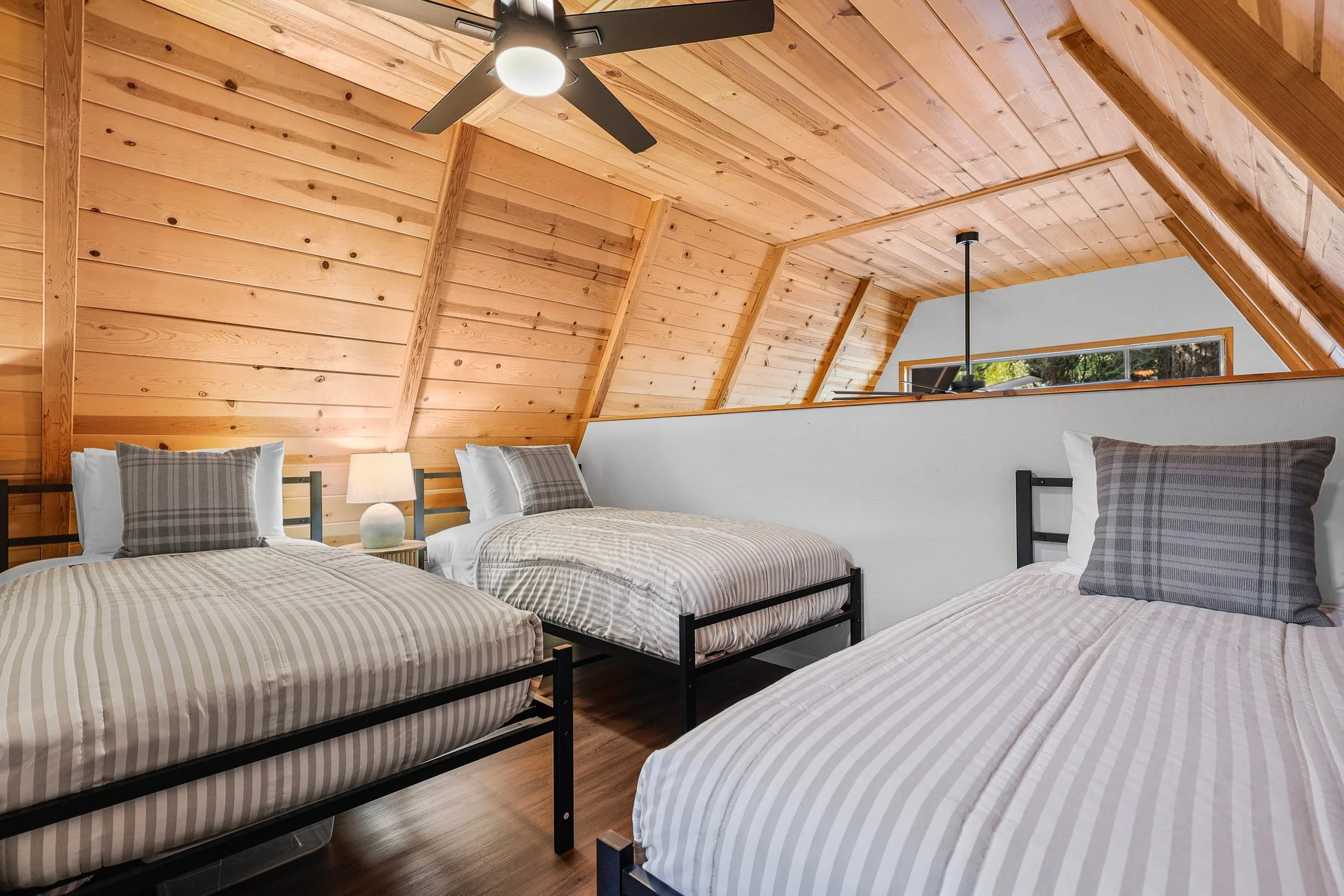 A bedroom with four twin beds with striped beige and white bedding, black metal frames, a bedside lamp, wood-paneled vaulted ceiling with a ceiling fan, and a window showing greenery outside.
