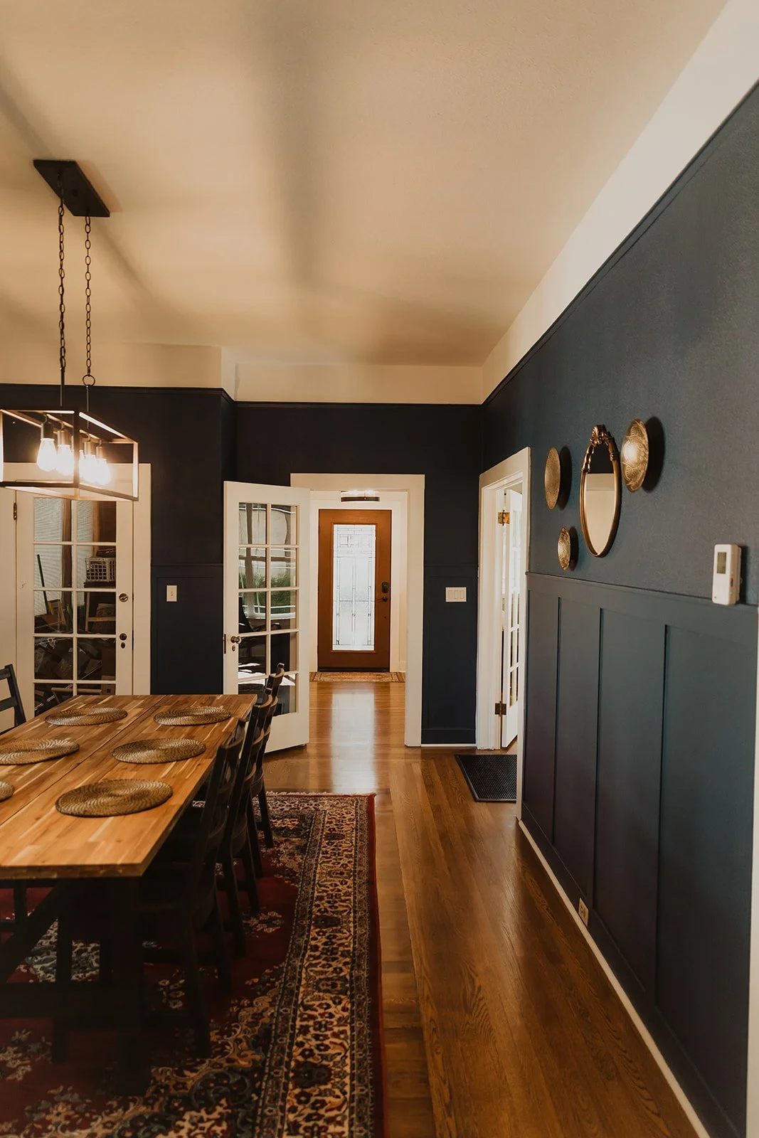 Dining room with a wooden table, wicker placemats, a patterned area rug, dark blue walls with decorative plates and mirror, and French doors leading to another room. Front door visible in the background.
