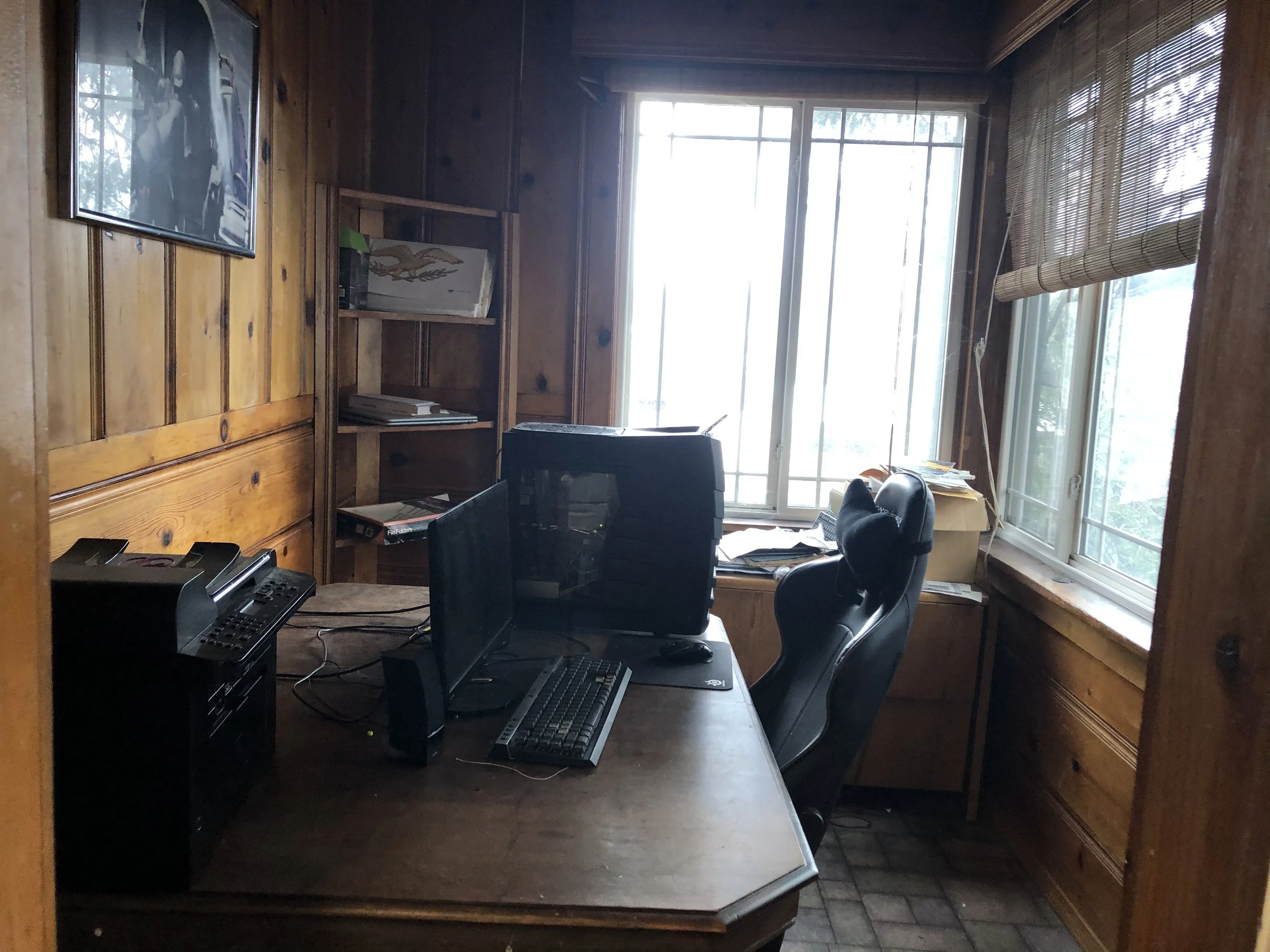 A home office with a wooden wall and large windows with blinds. There is a computer monitor, keyboard, and mouse on a desk, along with some papers. A black office chair is in front of the desk.