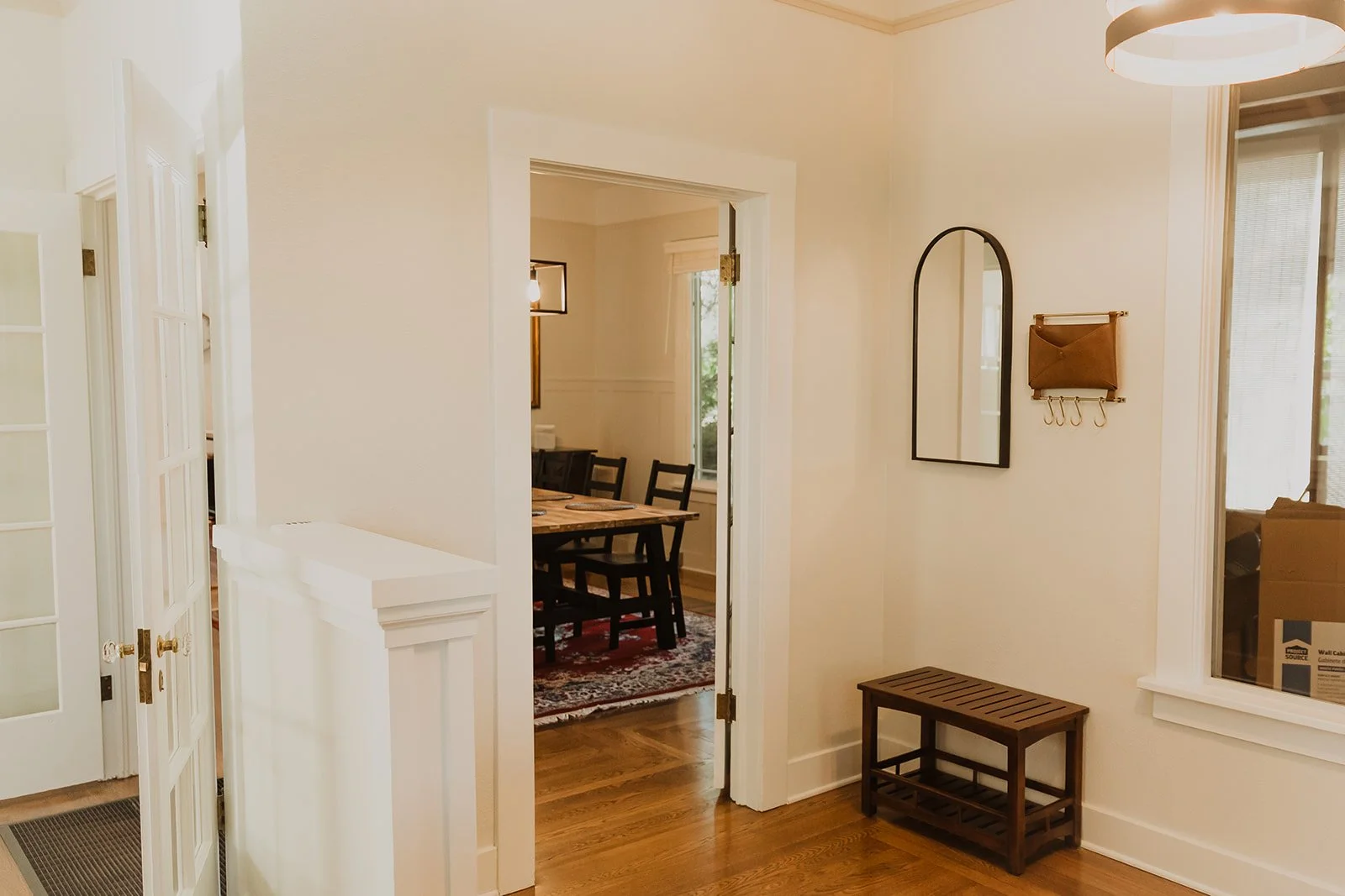 Interior view of a home with a hallway leading to a dining room. The hallway has a mirror, a wall-mounted bag holder with hooks, and a small bench. The dining room has a wooden table, black chairs, a window, and an area rug.