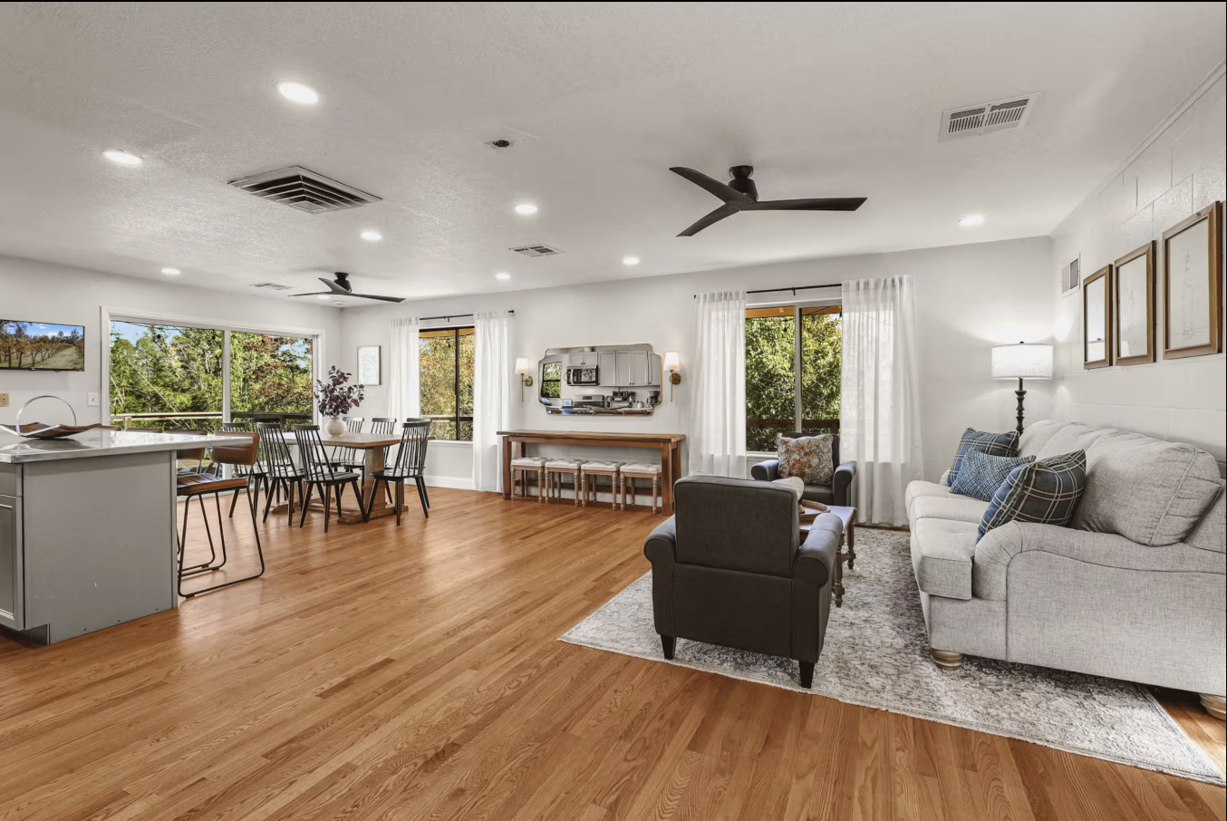 Living room with hardwood floors, white walls, large windows with white curtains, a gray sofa with blue pillows, a black armchair, a dining area with a round table and black chairs, and a kitchen counter visible on the left.