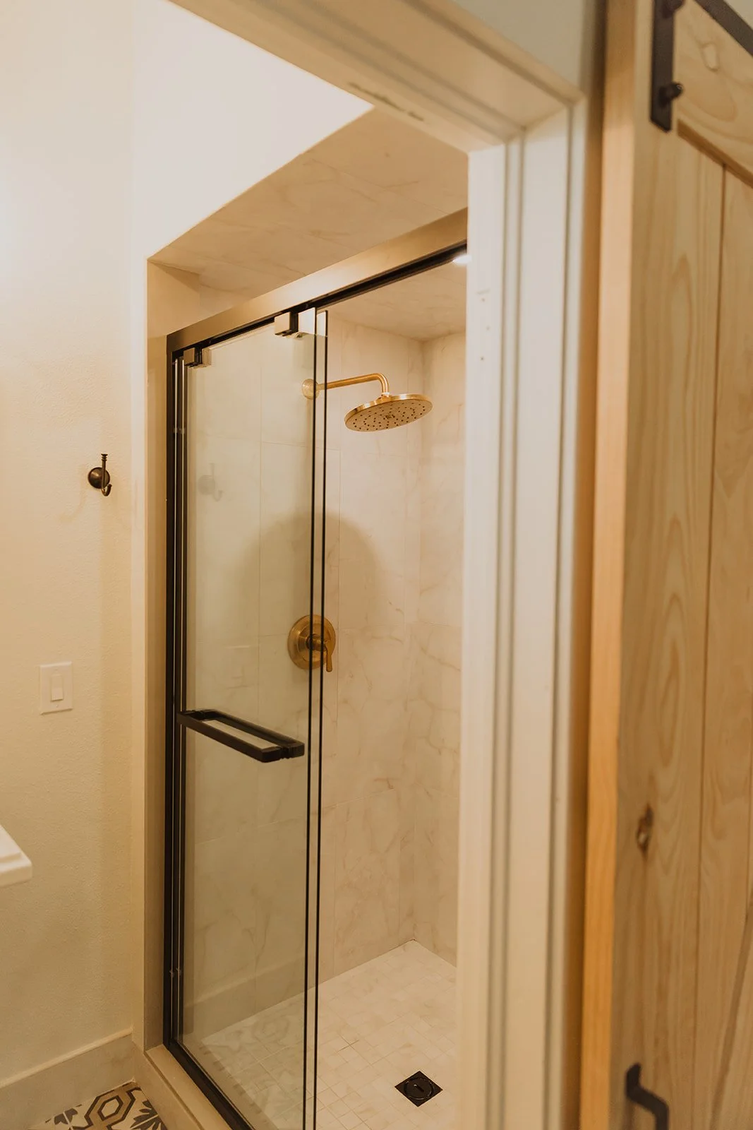 A small glass shower with a brass rain shower head and a black metal sliding door, set in a bathroom with beige and wooden accents.