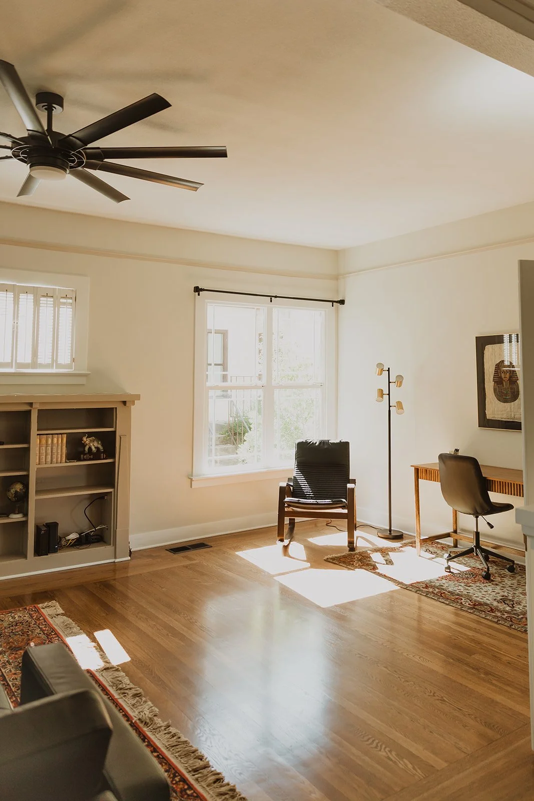 Bright living room with window, wooden floor, black chair, desk with office chair, standing lamp, bookshelf, artwork, and ceiling fan.