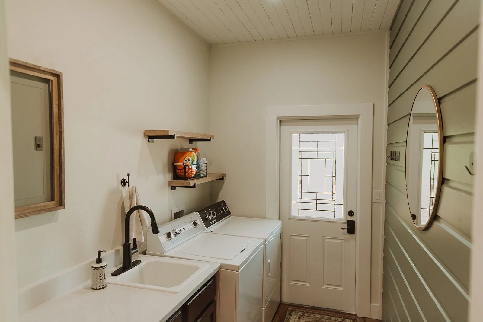 A laundry room with a white sink, a black faucet, a soap dispenser, a washer and dryer, wooden wall shelves with laundry supplies, a mirror, a window with a stained glass design, and a door with a window and stained glass