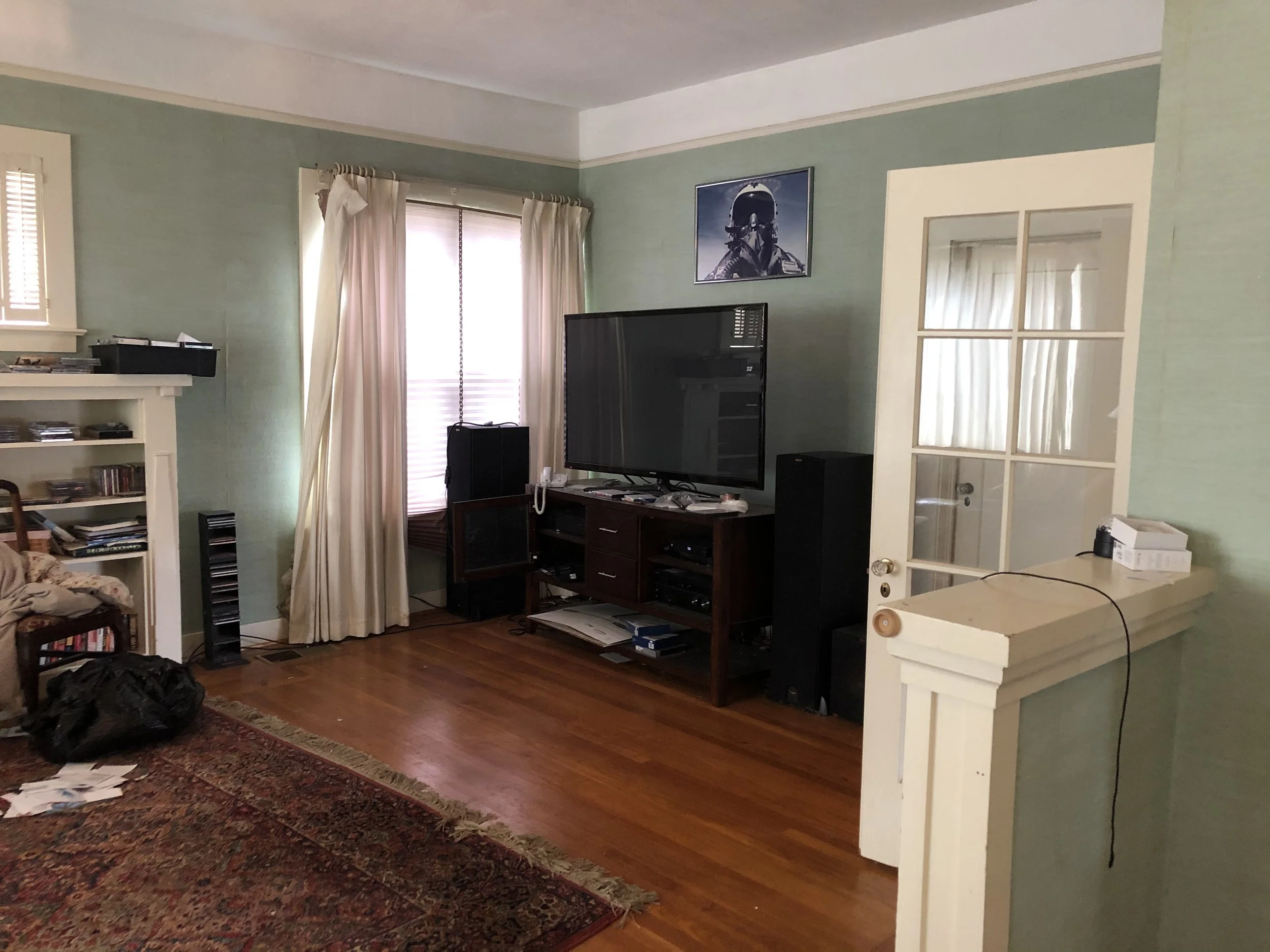 A living room with green walls, hardwood flooring, a window with curtains, a flat-screen TV on a wooden stand, and an open white door with glass panes.