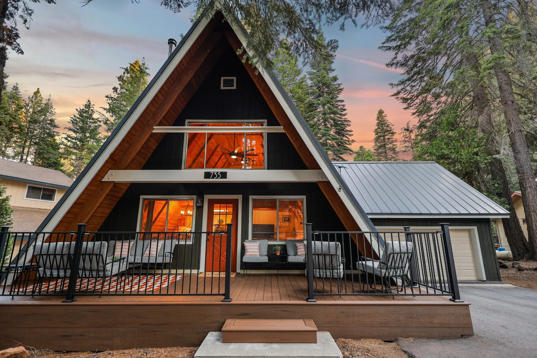 A modern A-frame house with large glass windows and a wooden deck, surrounded by tall trees at sunset.